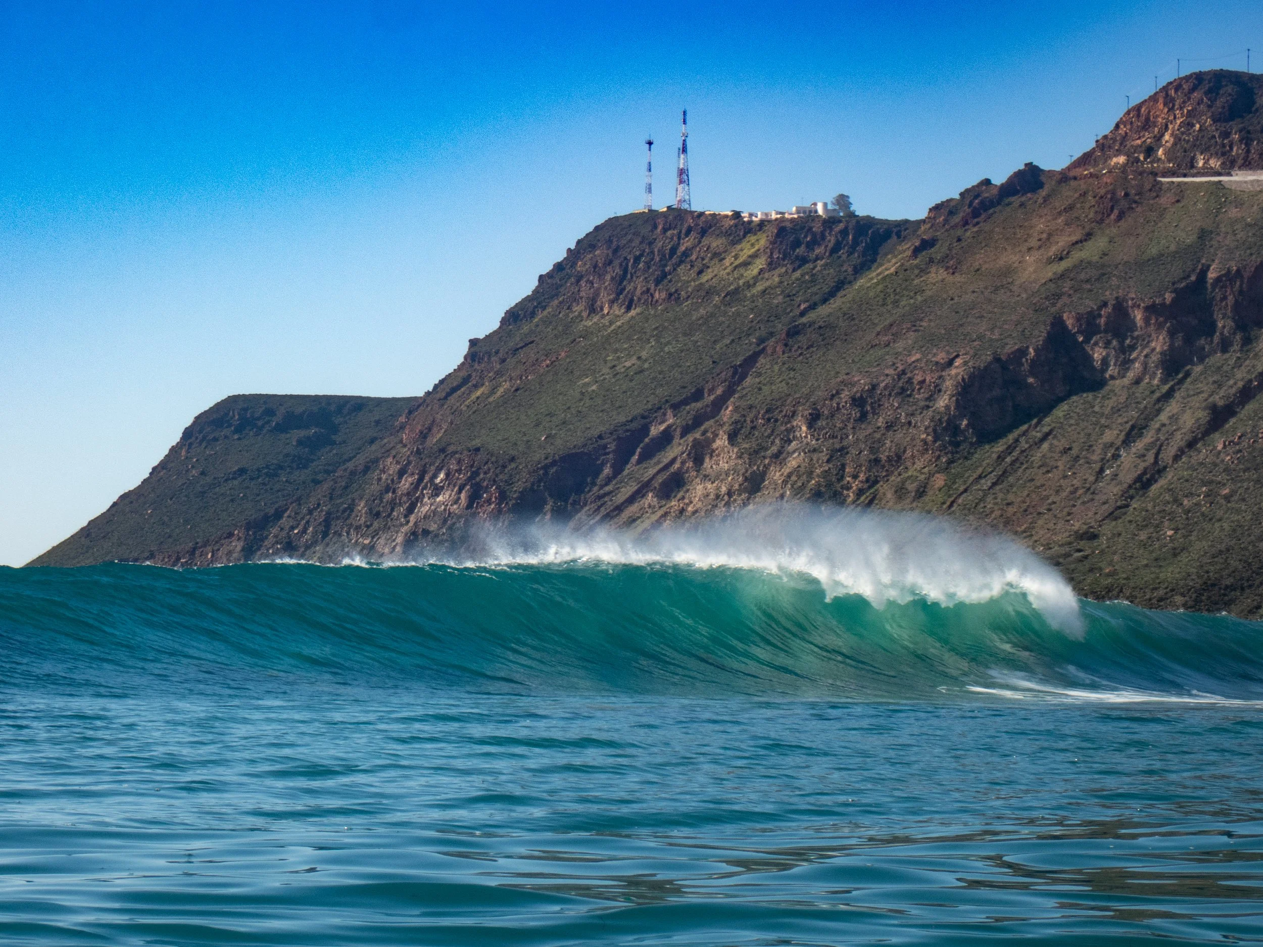 Large ocean wave breaking near a rugged coastline with green hills and communication towers on top, under a clear blue sky.