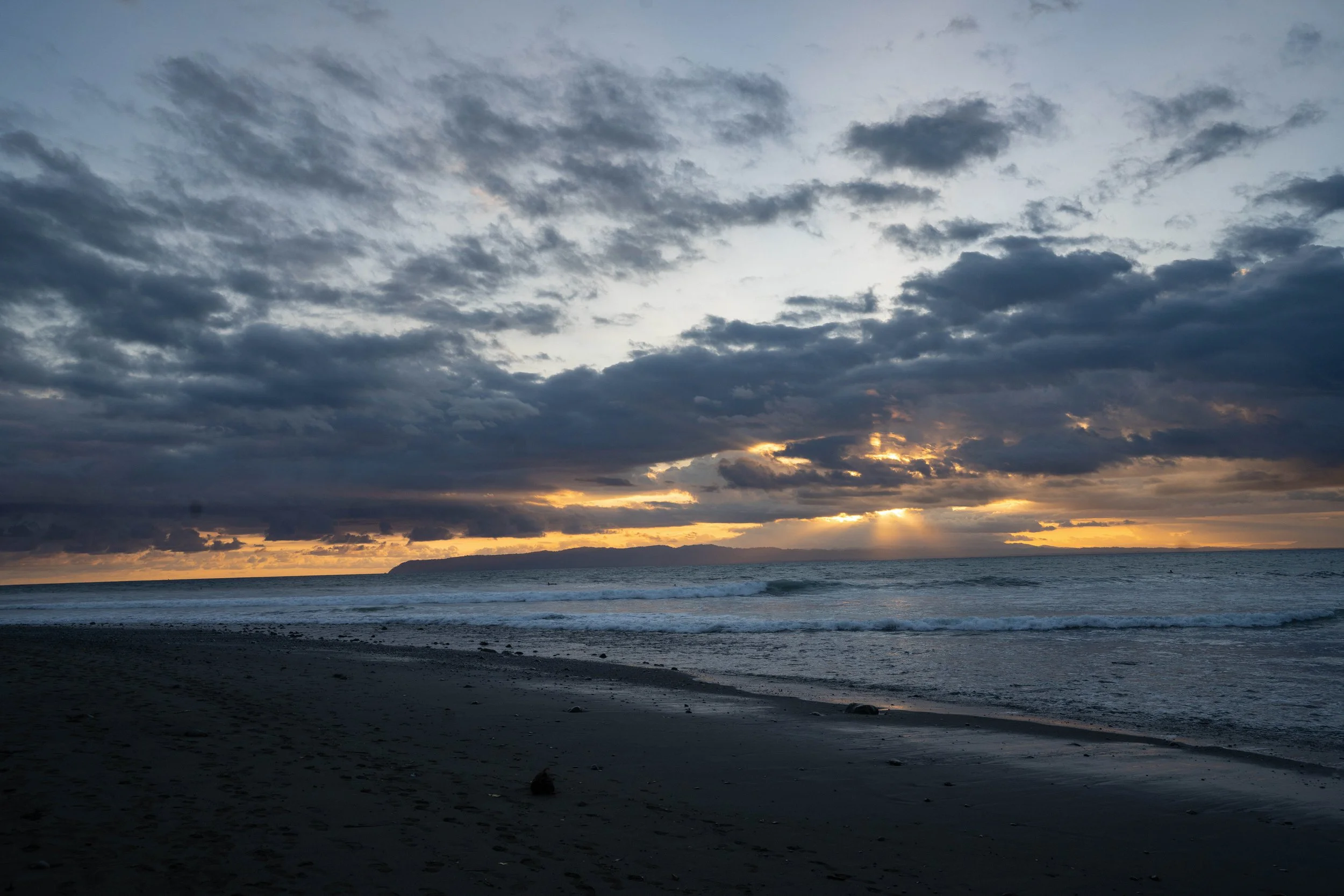 A view of a cloudy sky during sunset over a calm ocean with dark sand beach in foreground.