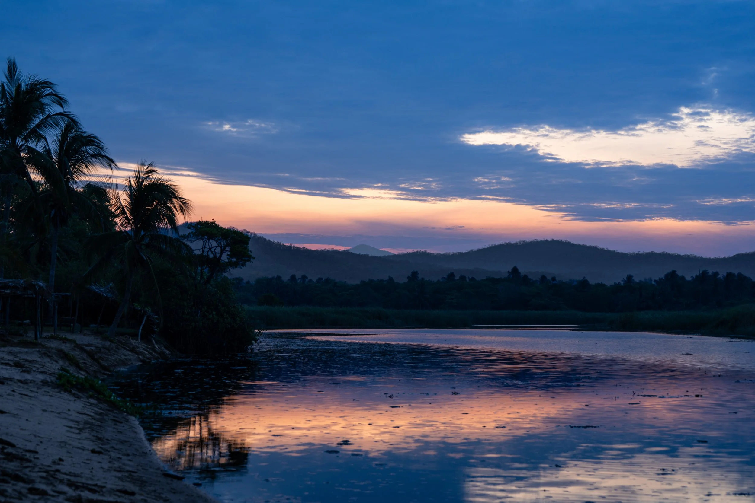 Sunset over a river with silhouetted trees and distant hills, reflecting colorful sky in the water.