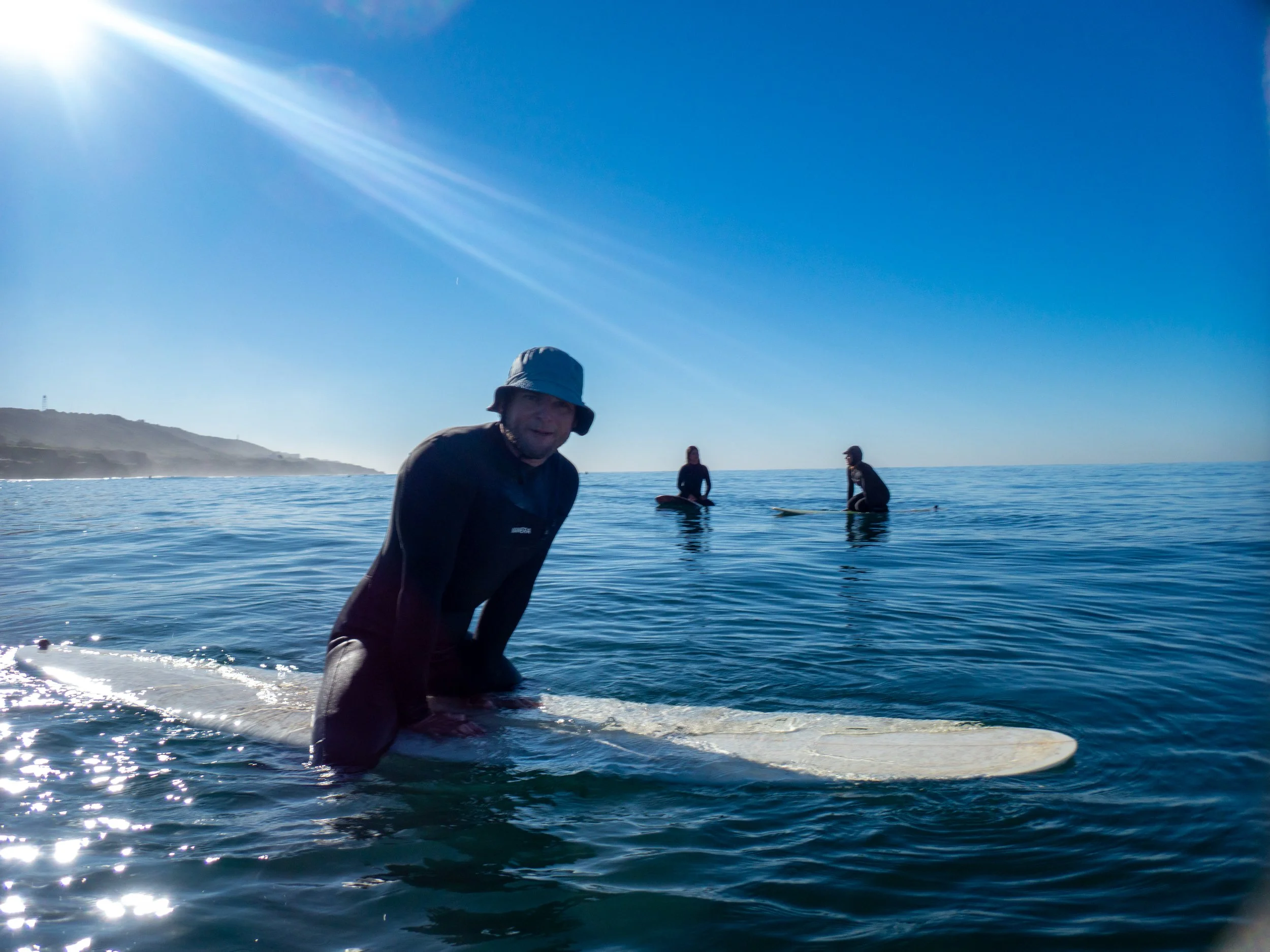 A man kneeling on a surfboard in the ocean with two others in the background, all preparing to surf under a clear blue sky with the sun shining brightly.