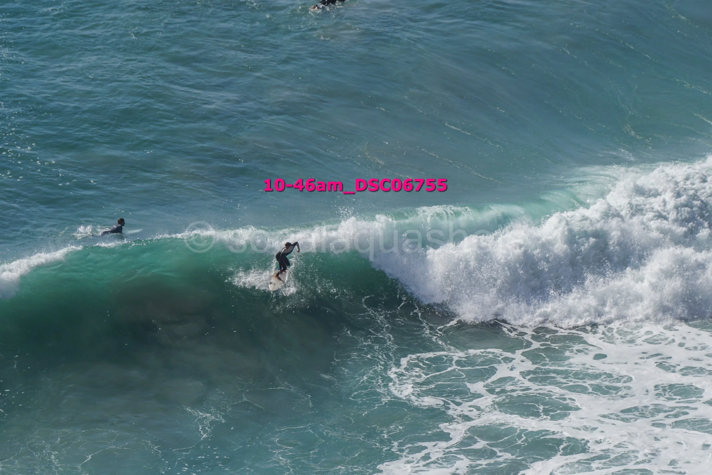 A person surfing on a wave in the ocean with another person in the background, with pink text overlay reading '10-46am_DS06755'.