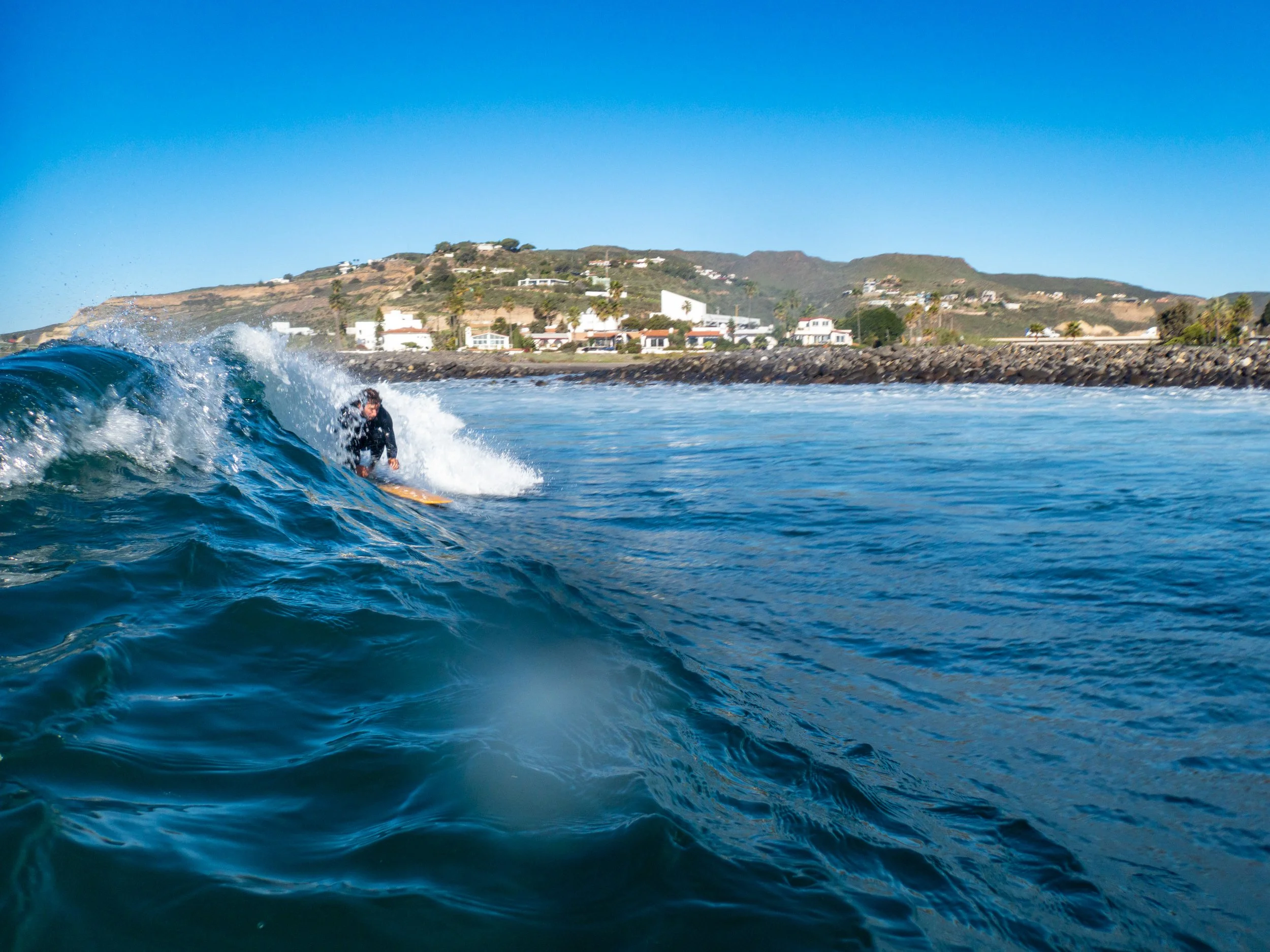 Person surfing on a wave with a coastal houses and hills in the background.