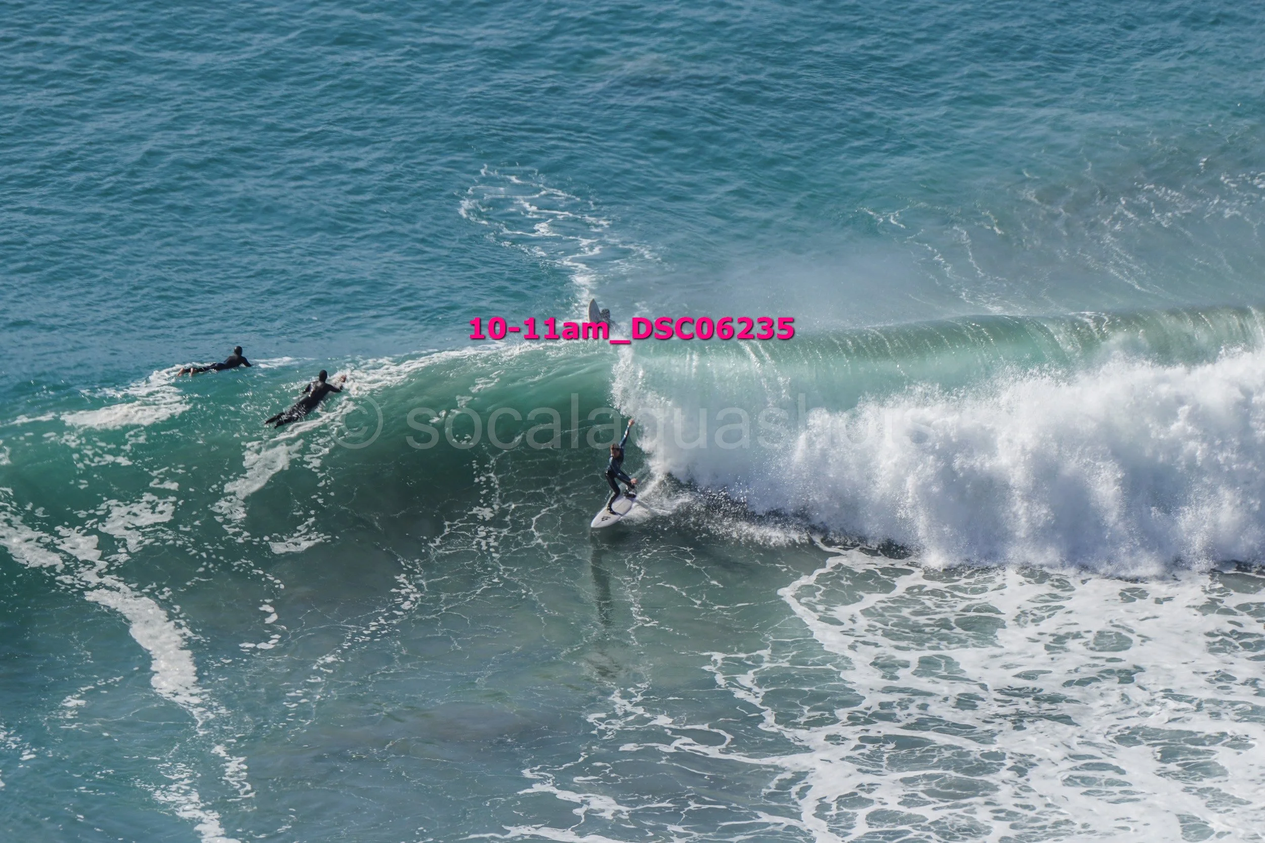 A person surfing on a large wave with two other surfers paddling in the water nearby.