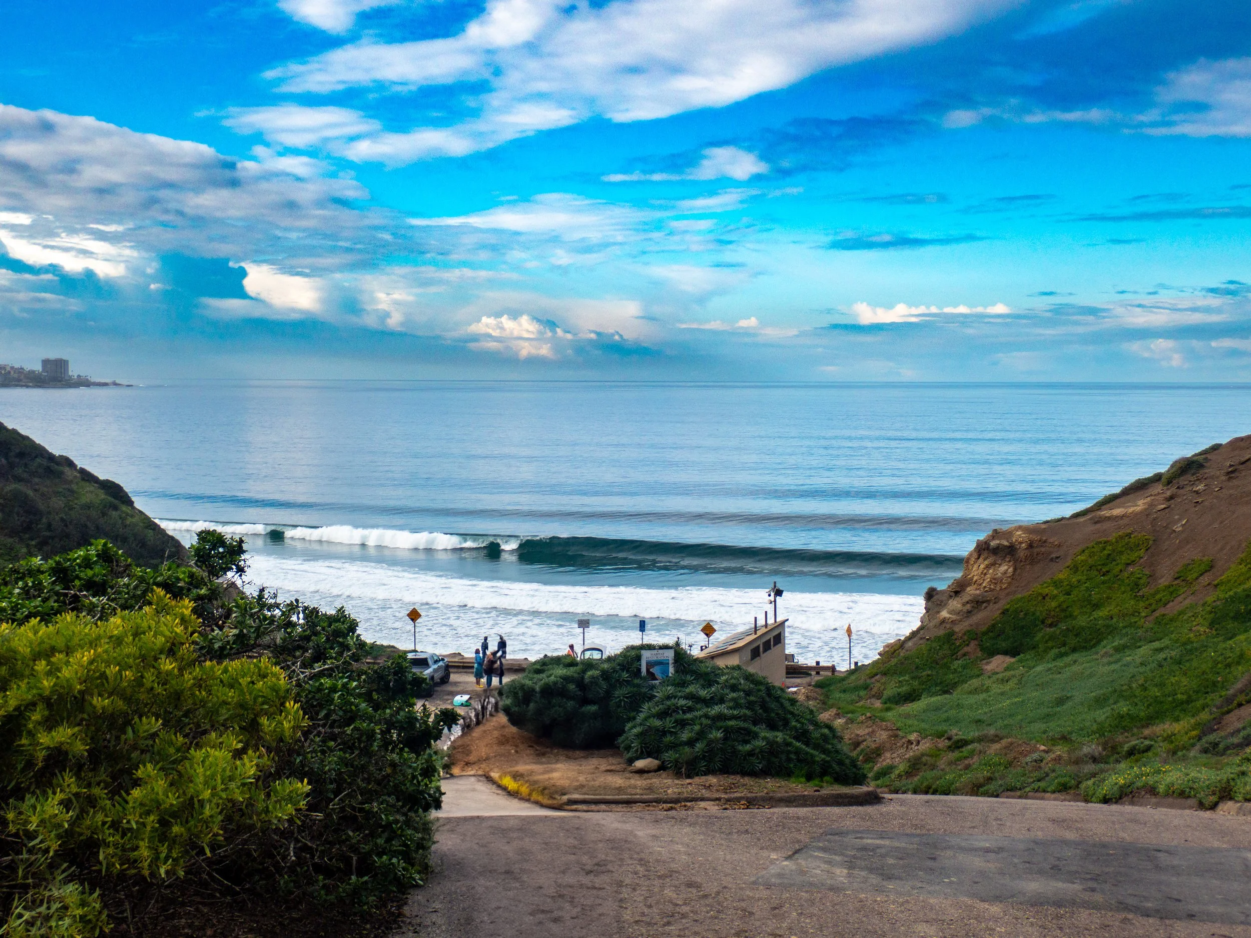 View of the ocean with waves, partly cloudy sky, green bushes and hills framing the scene, and a pathway leading to the beach.