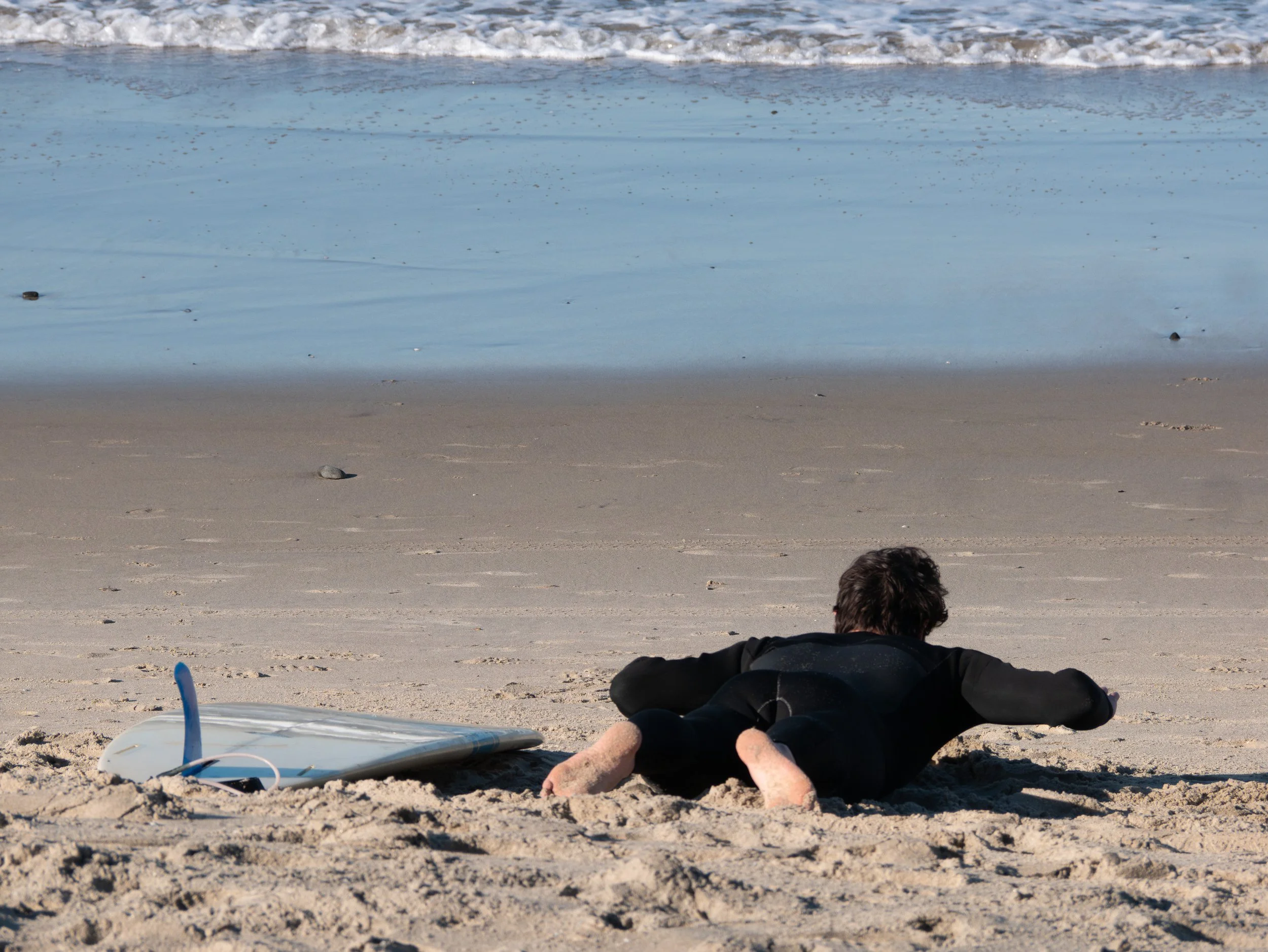 Person lying on the sandy beach near a surfboard, facing the ocean with small waves in the background.