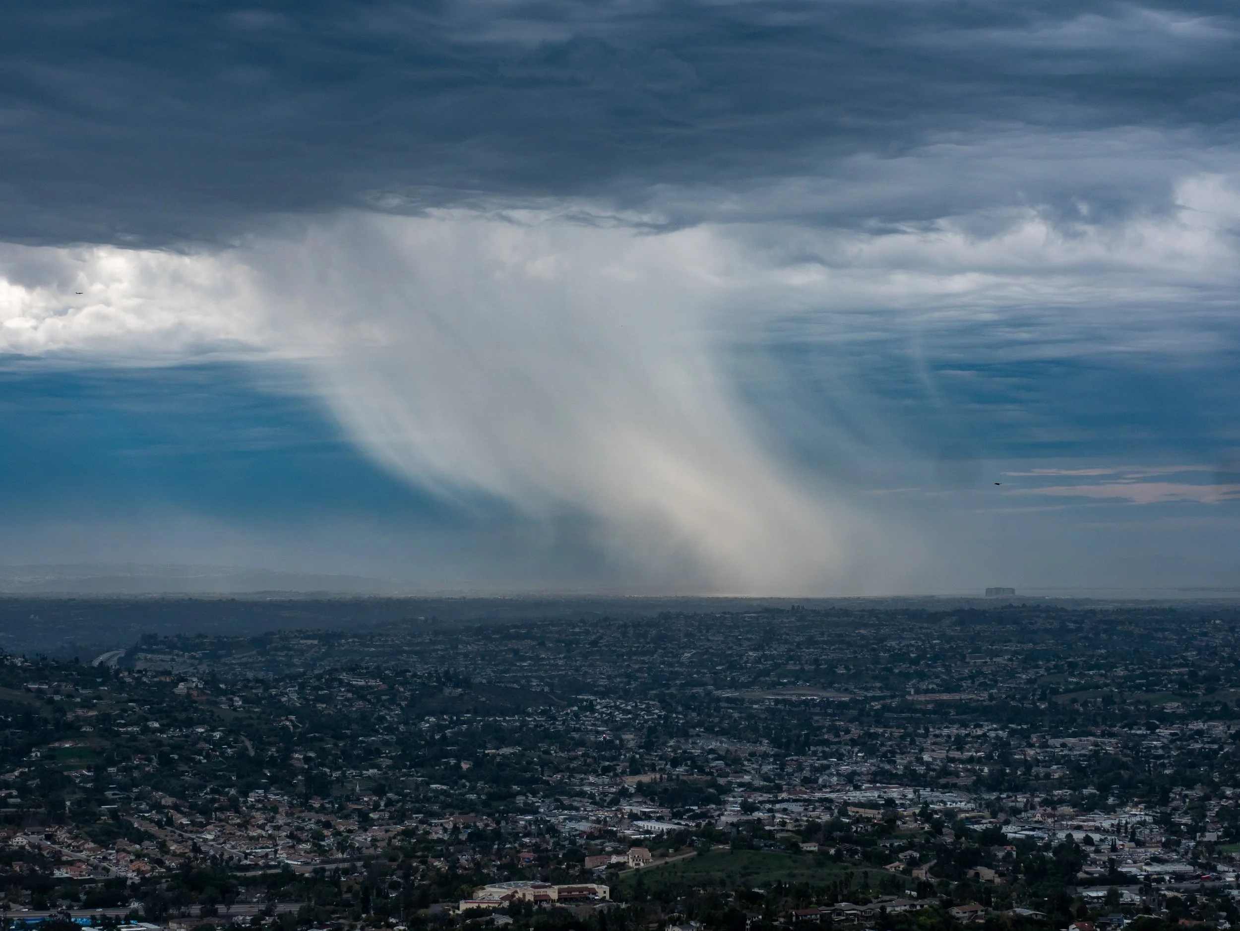 A cityscape under dark, stormy clouds with a rain shaft descending from the clouds to the ground.