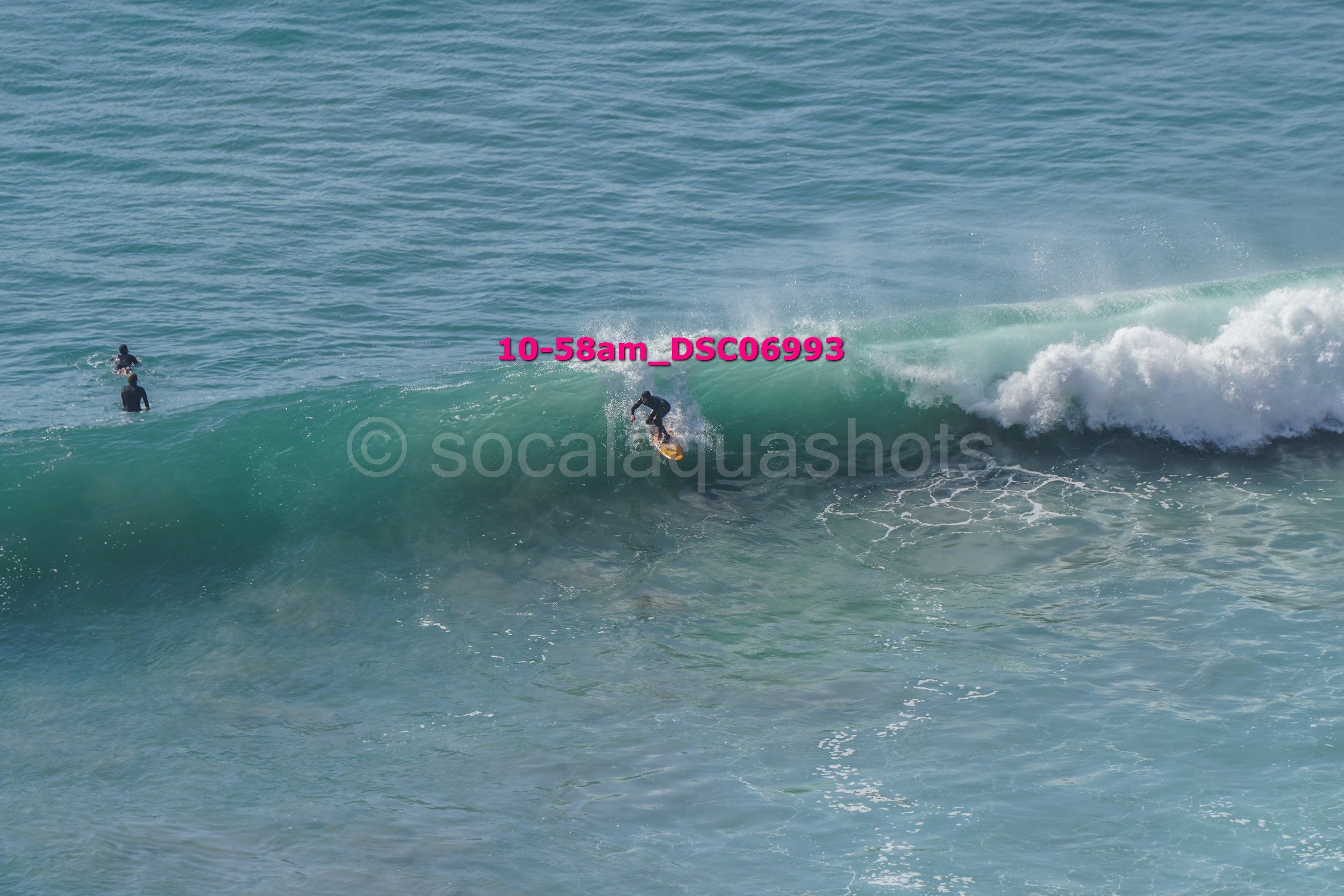 Surfer riding a wave with two people in the water nearby, ocean water and foam in the background.