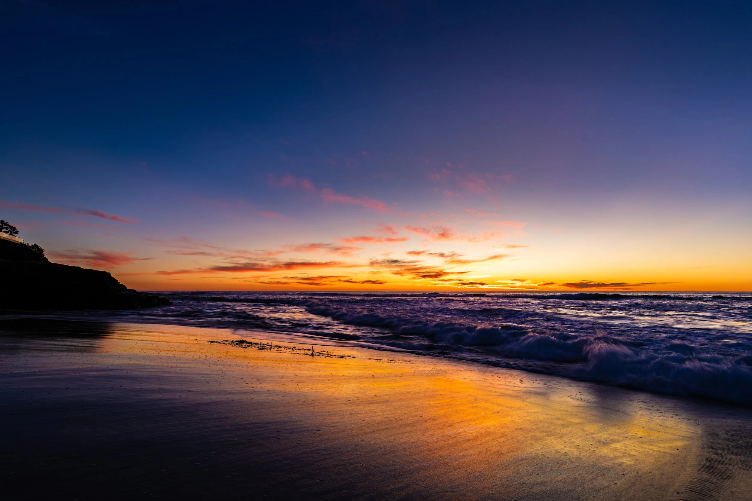A colorful sunset over the ocean, with waves crashing onto the sandy beach and a silhouette of a hill on the left.