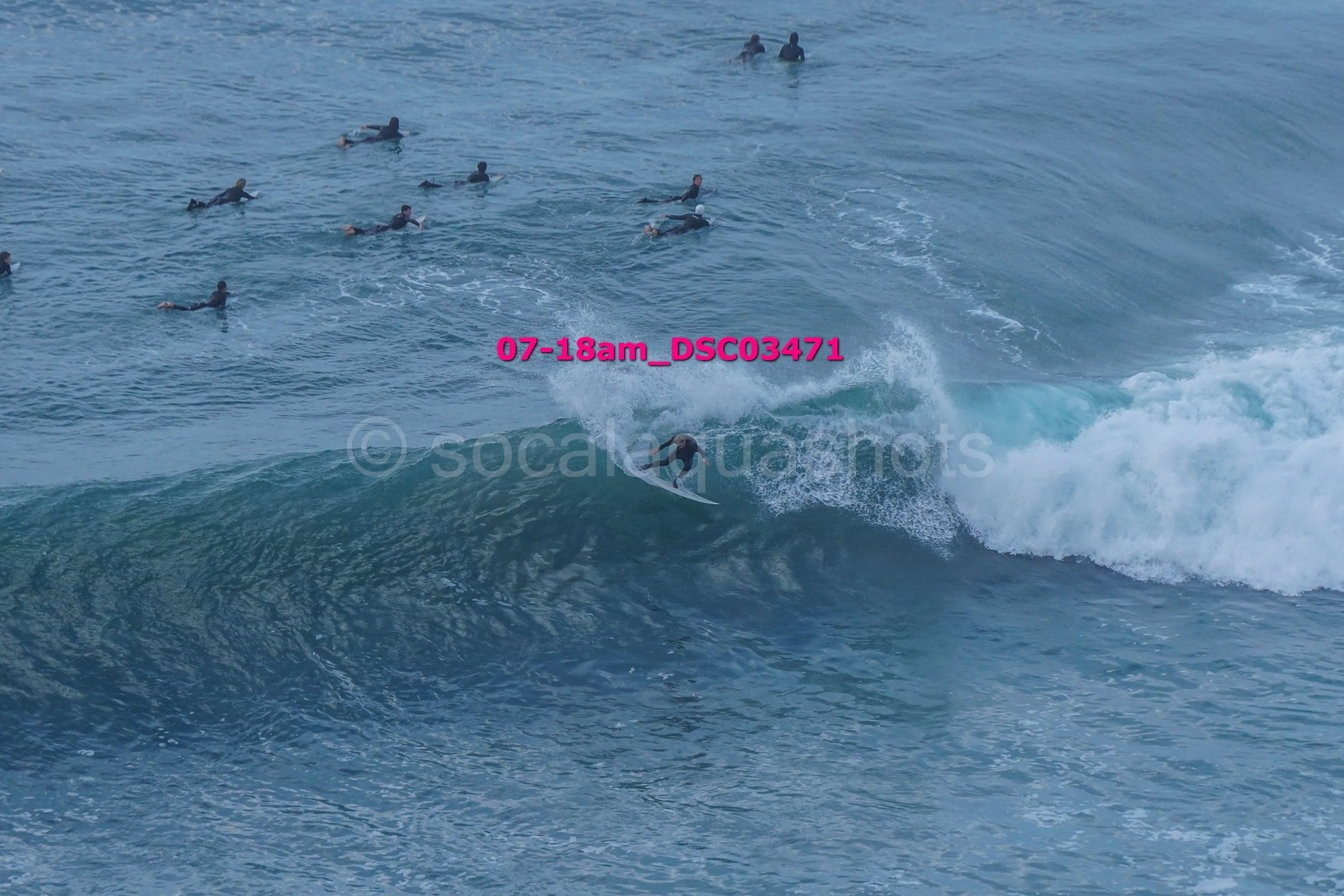 Surfer riding a wave while multiple people swim or surf in the ocean nearby.
