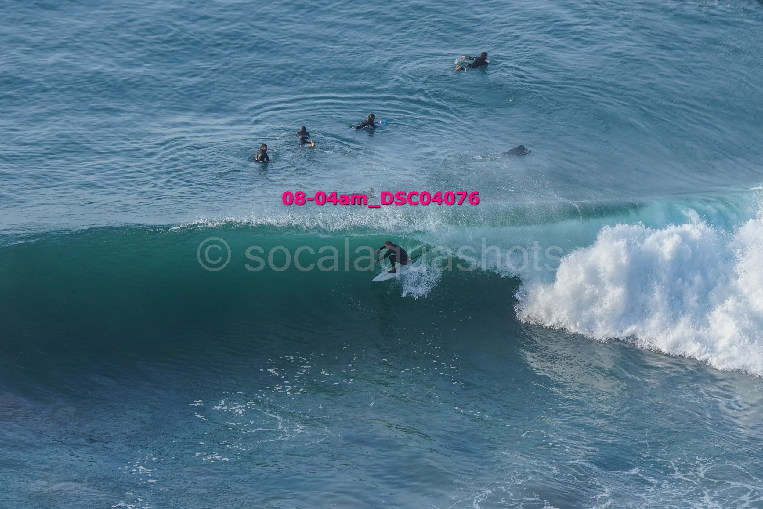 Surfer riding a large wave with several people swimming in the ocean background.