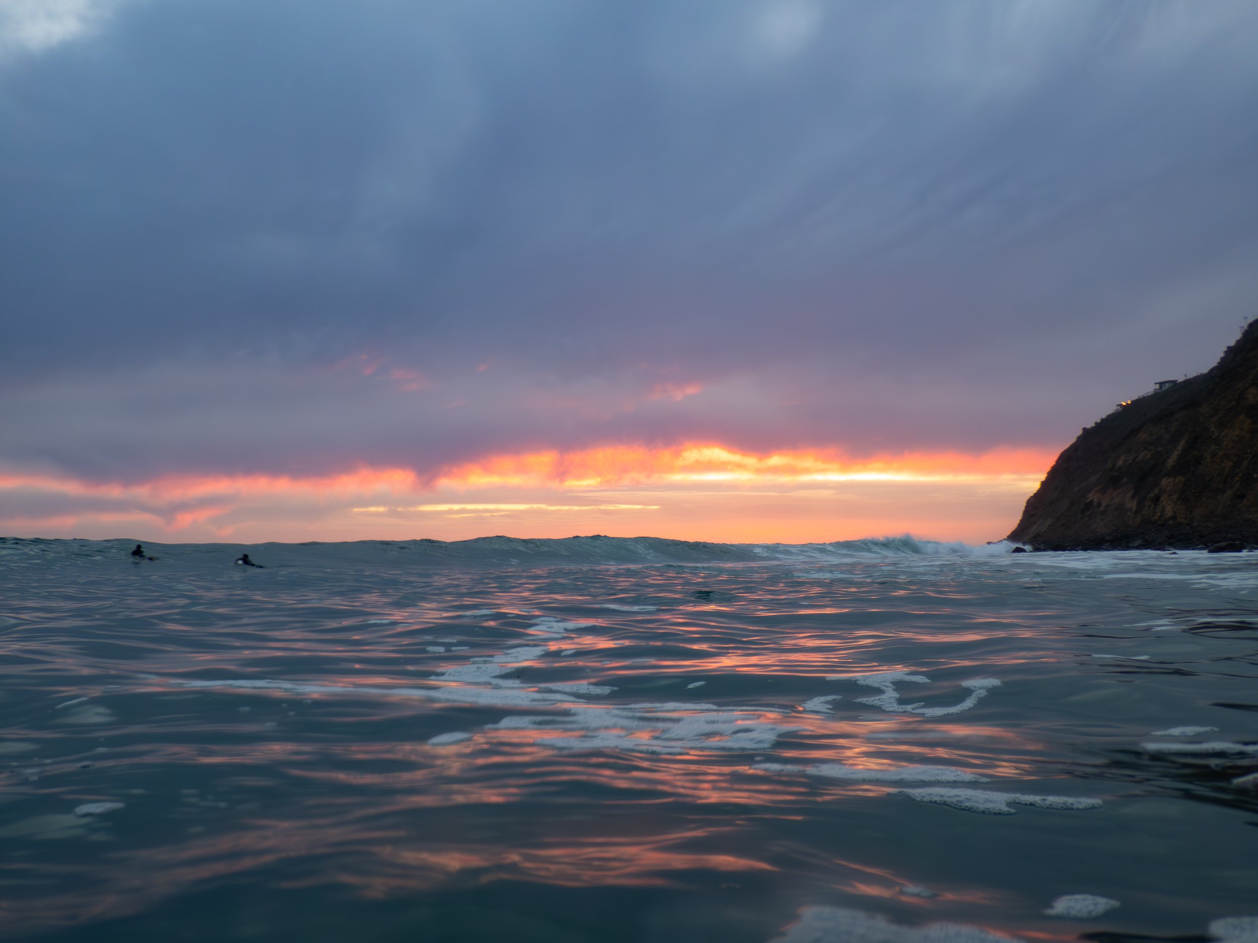 Ocean at sunset with waves and a cloudy sky, a cliffside on the right, and two surfers in the water.