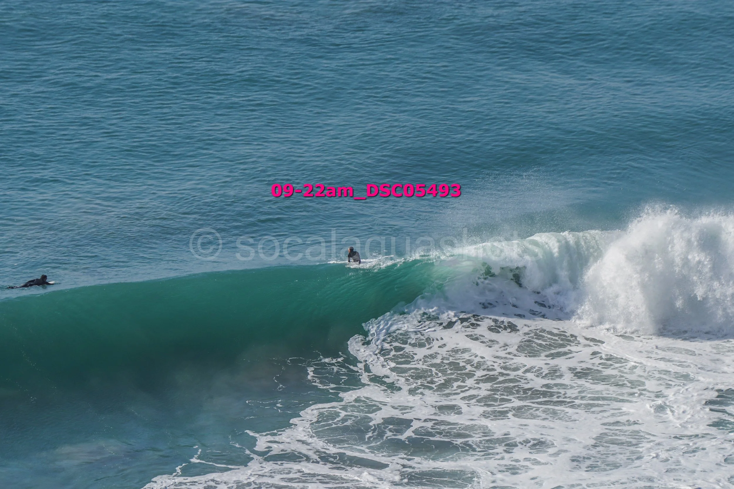 Two surfers in wetsuits riding a large ocean wave with white foam at the crest, ocean water in the background.