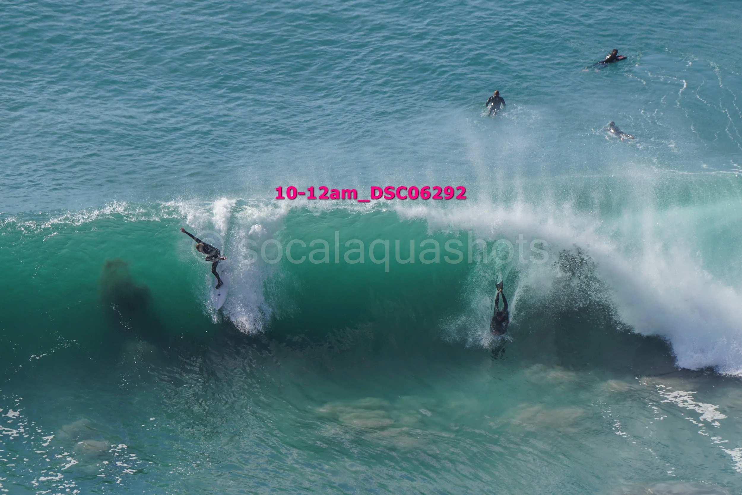 Surfers riding a large wave with four other surfers swimming nearby in the water.