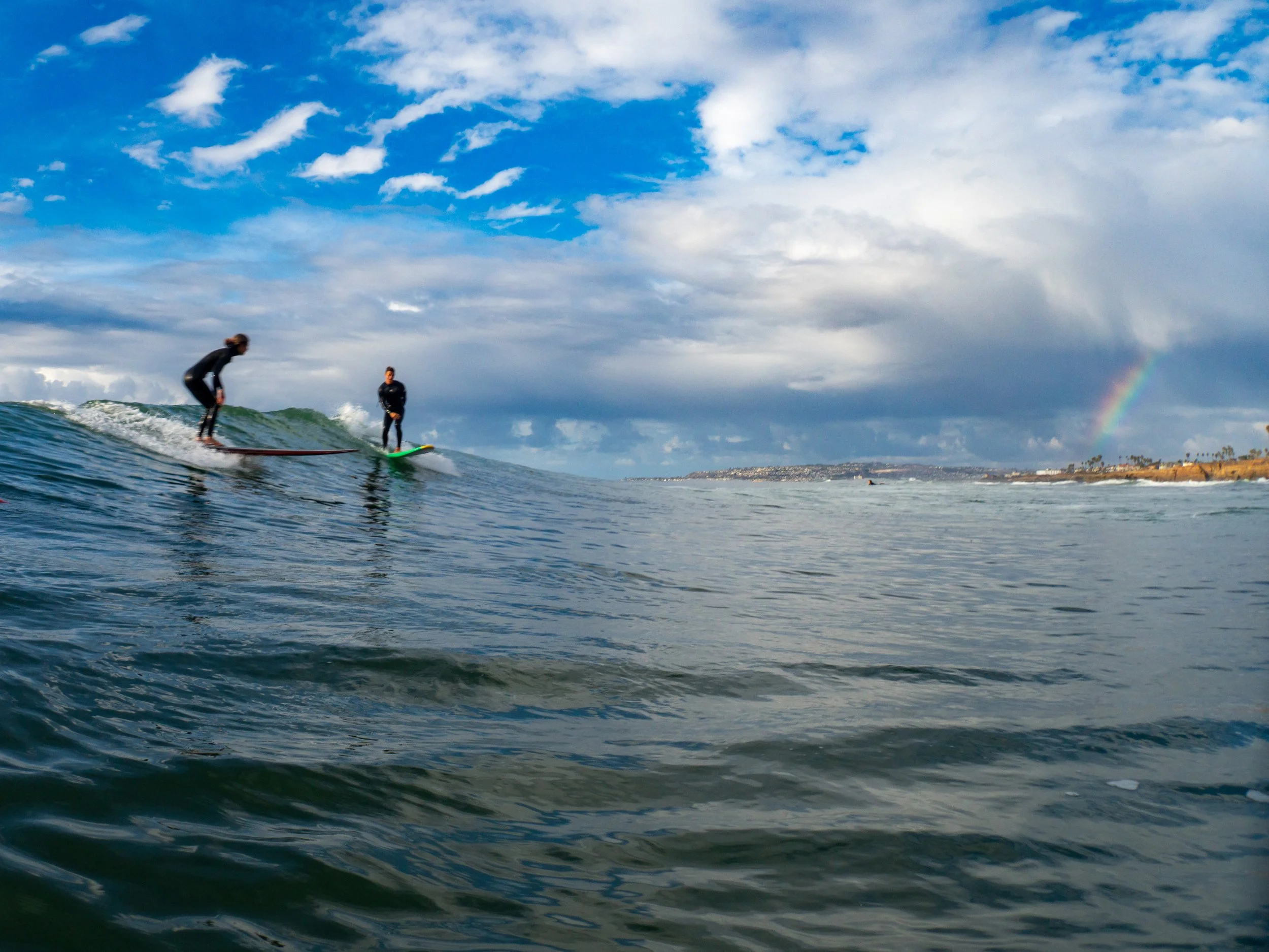 Two people surfing on the ocean with a rainbow in the background and a partly cloudy sky.