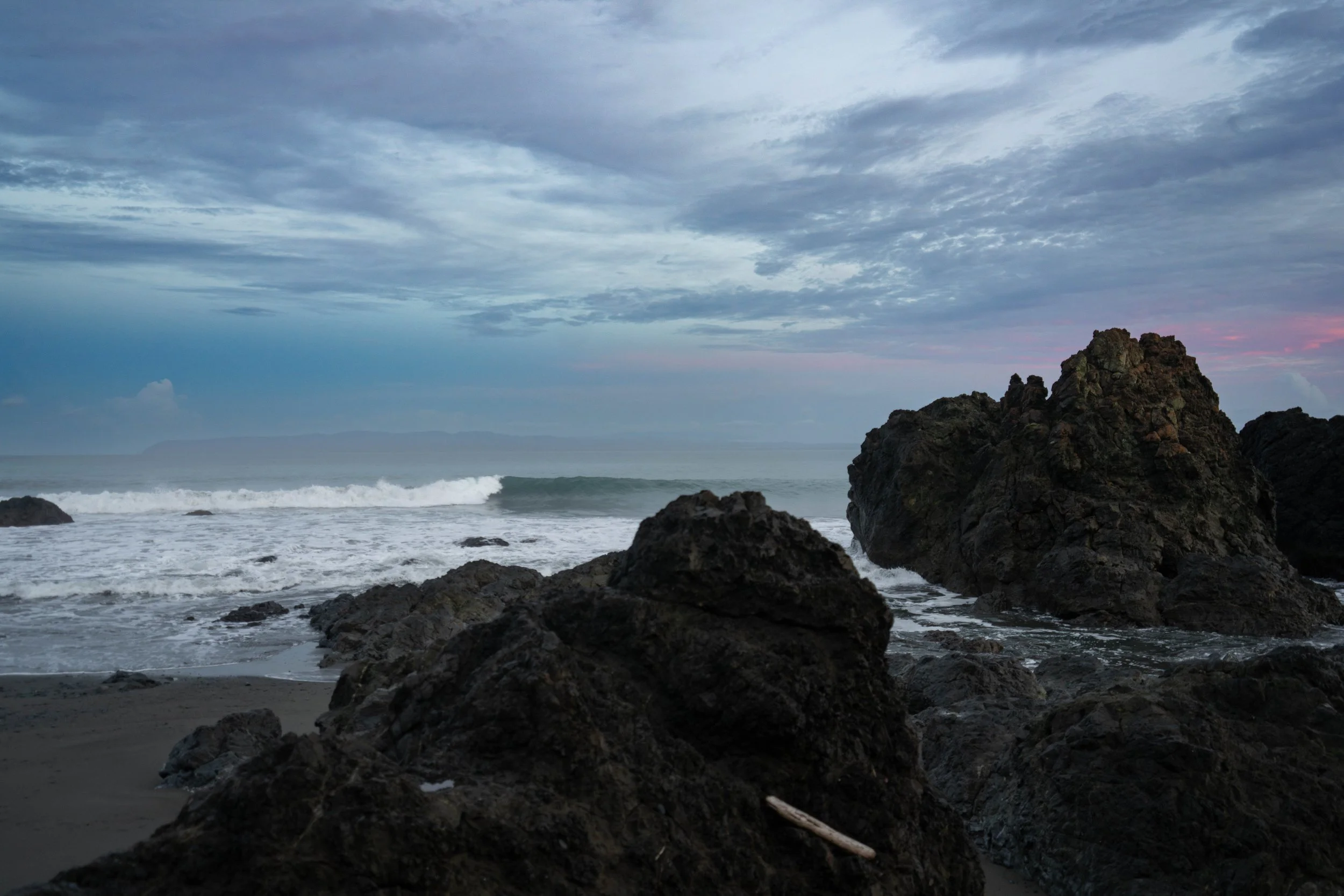 A rocky coastline with dark rocks in the foreground, waves crashing on the shore, and an overcast sky with blue and gray clouds.
