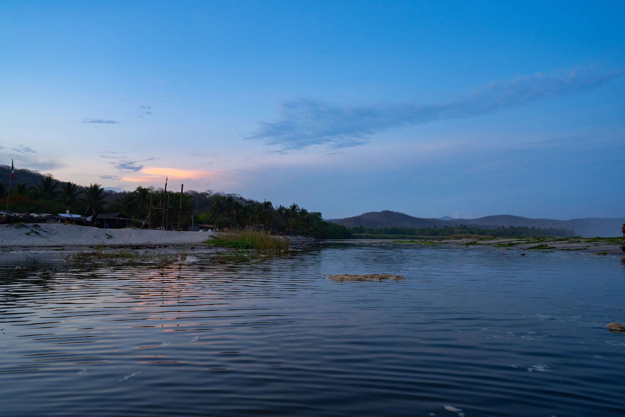 A river or lake at dusk with calm water reflecting the blue sky, some clouds, distant hills, and a shoreline with palm trees and small structures.