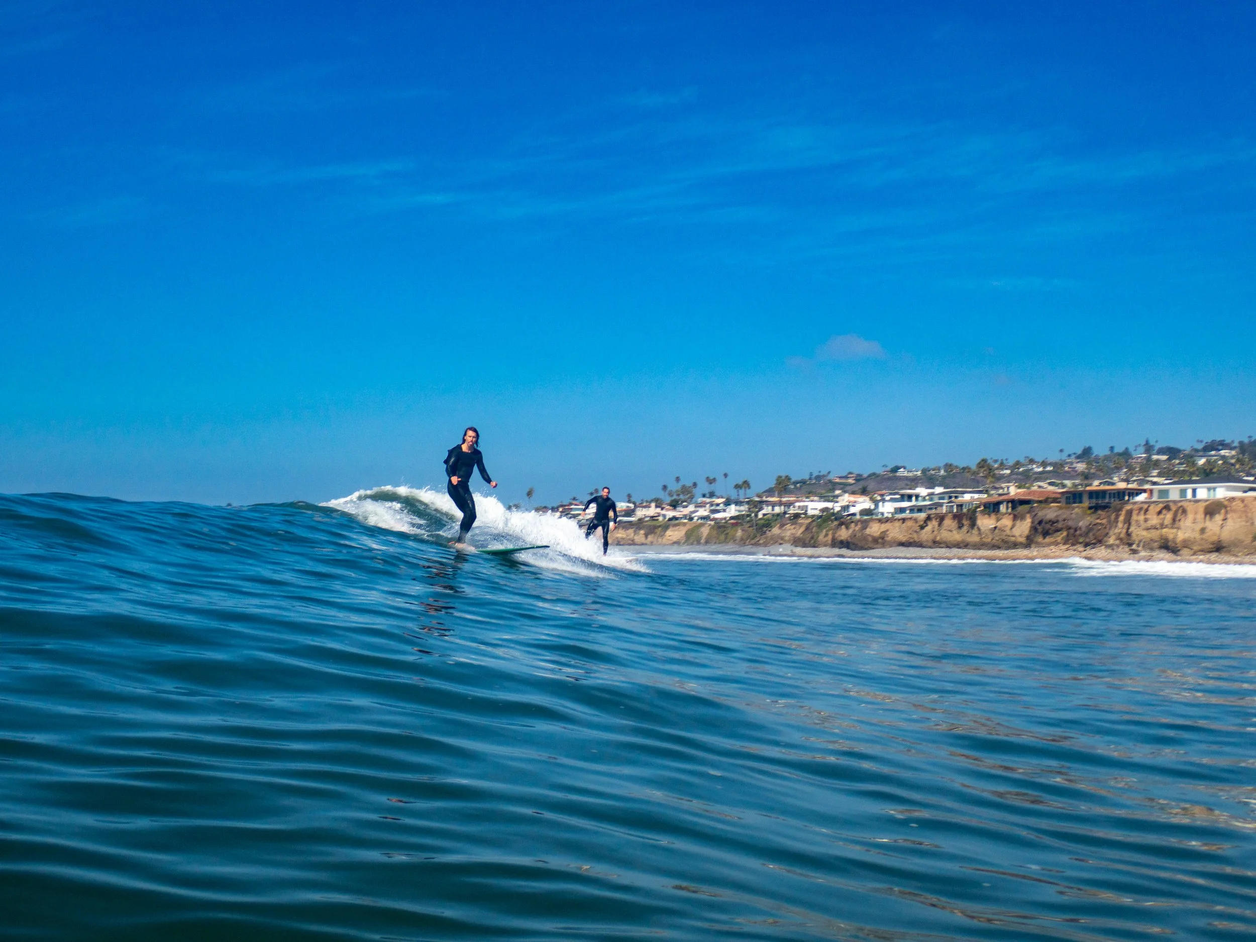 Two women surfing on the ocean with a shoreline and houses in the background under a blue sky.