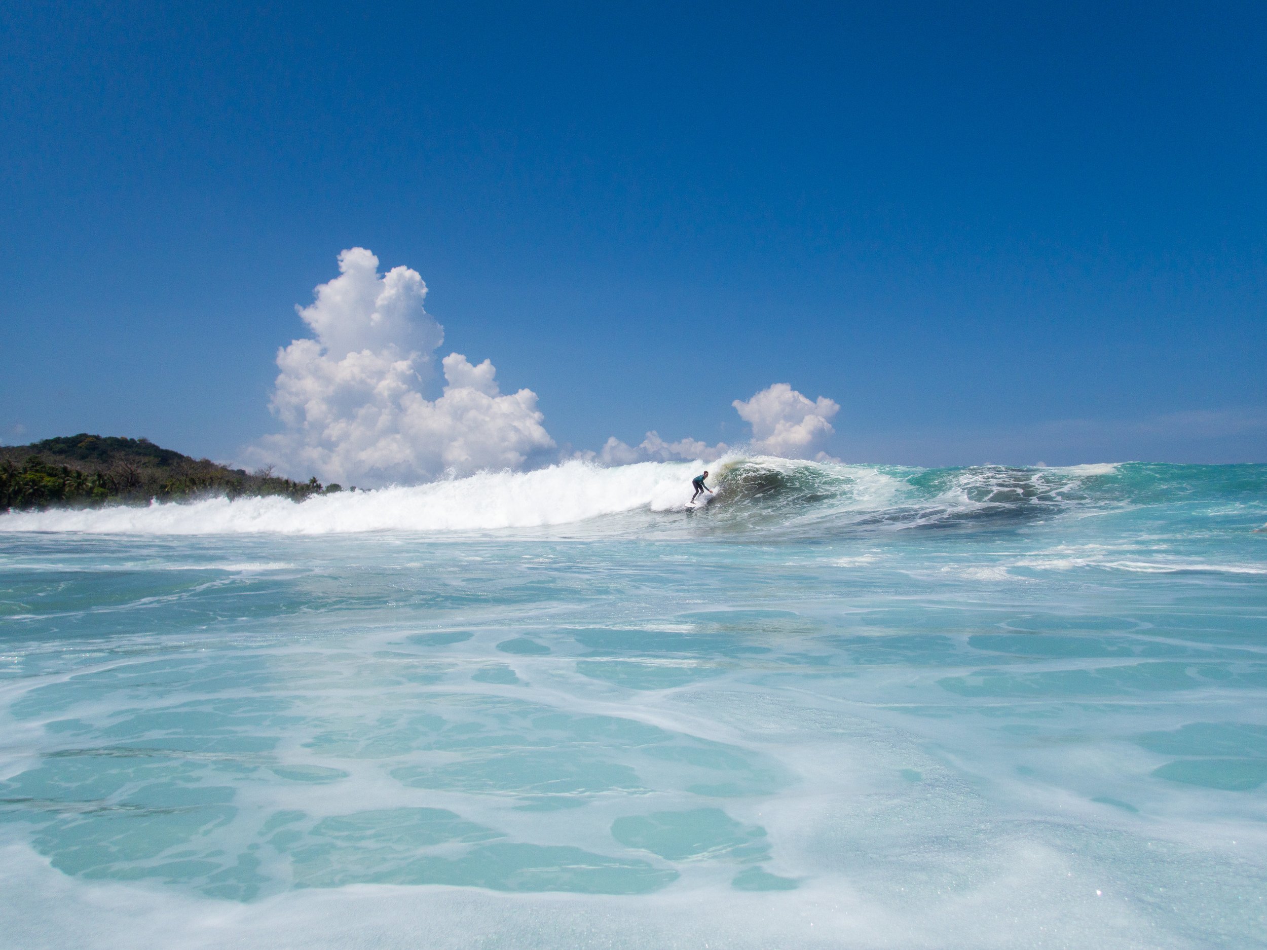 Surfer riding a wave in clear blue ocean under a bright blue sky with white clouds.