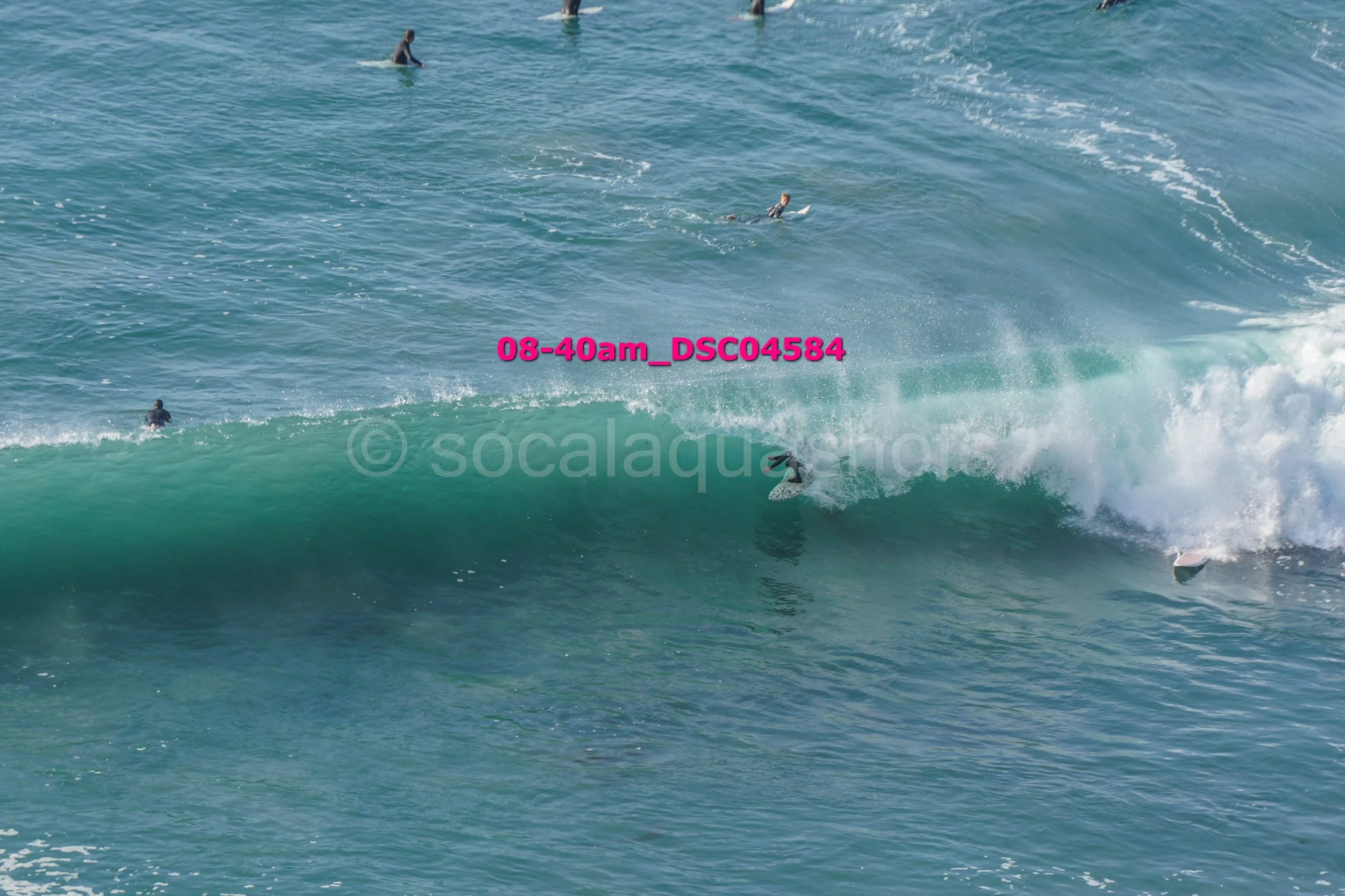 People surfing in the ocean with waves. One surfer is riding a wave, others are swimming or waiting in the water.