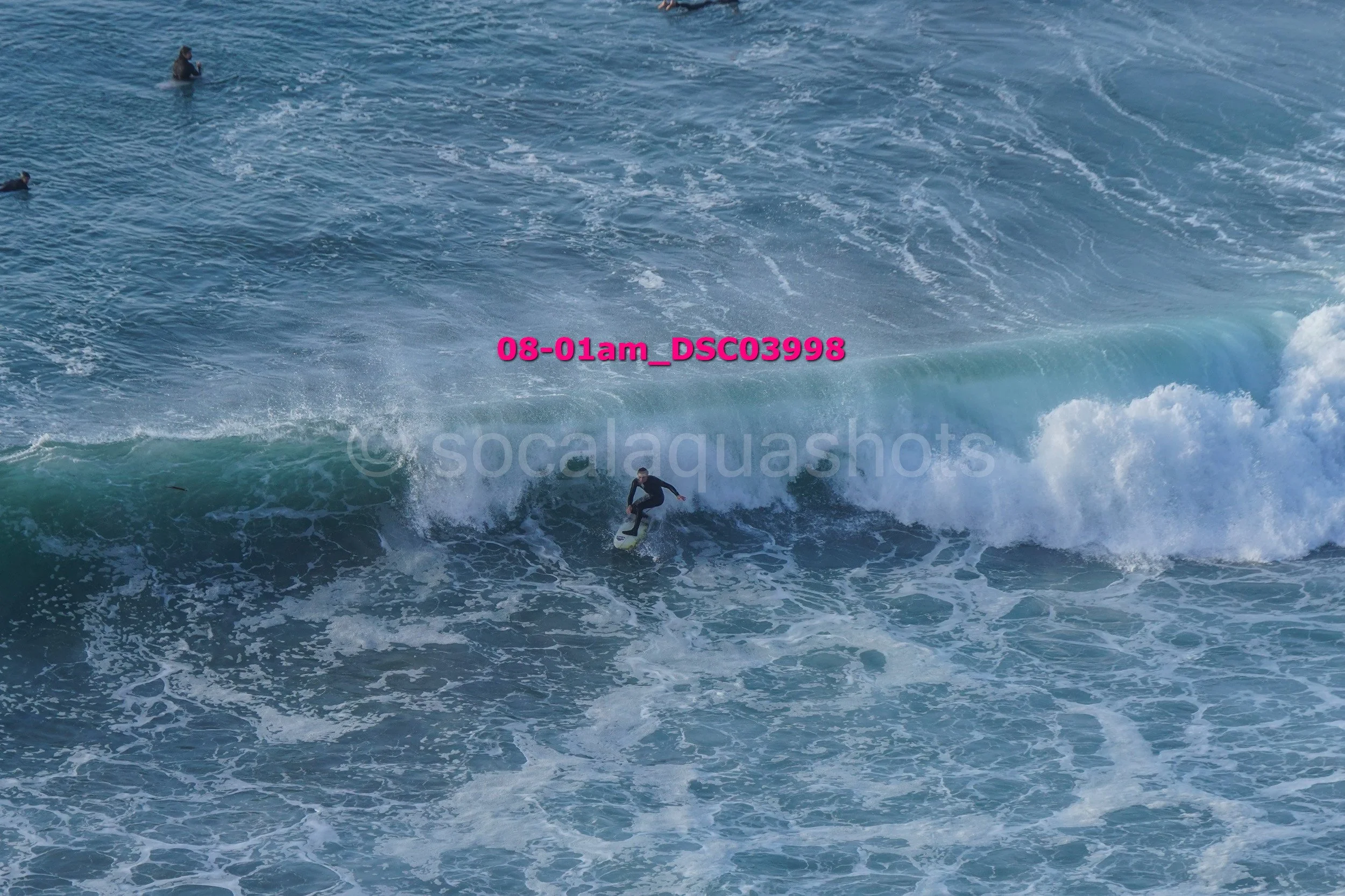 A person surfing on a wave in the ocean, surrounded by smaller waves and water. There are a few other people in the water nearby.