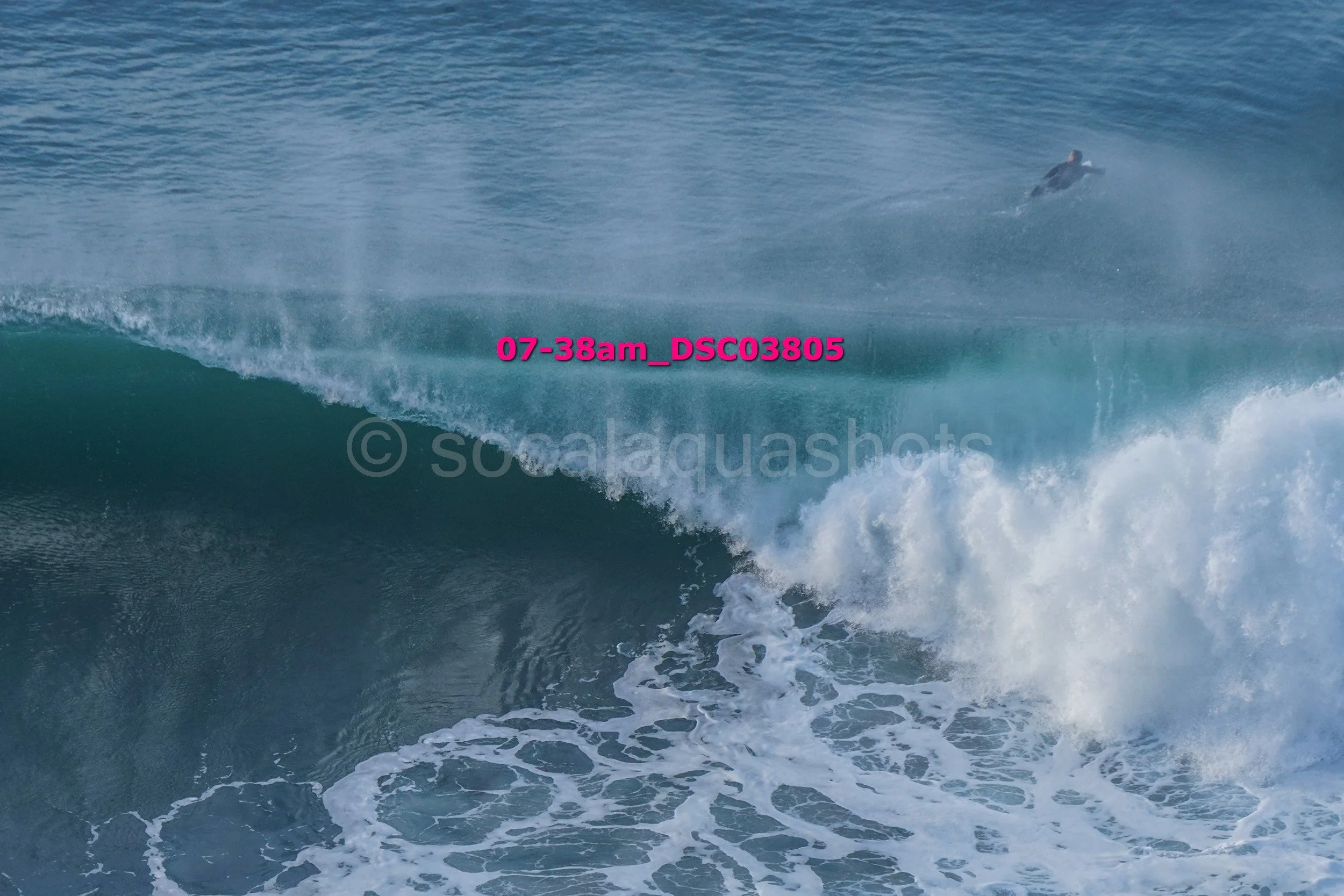 A surfer riding a large ocean wave with spray and white foam, deep blue water, and sky in the background.