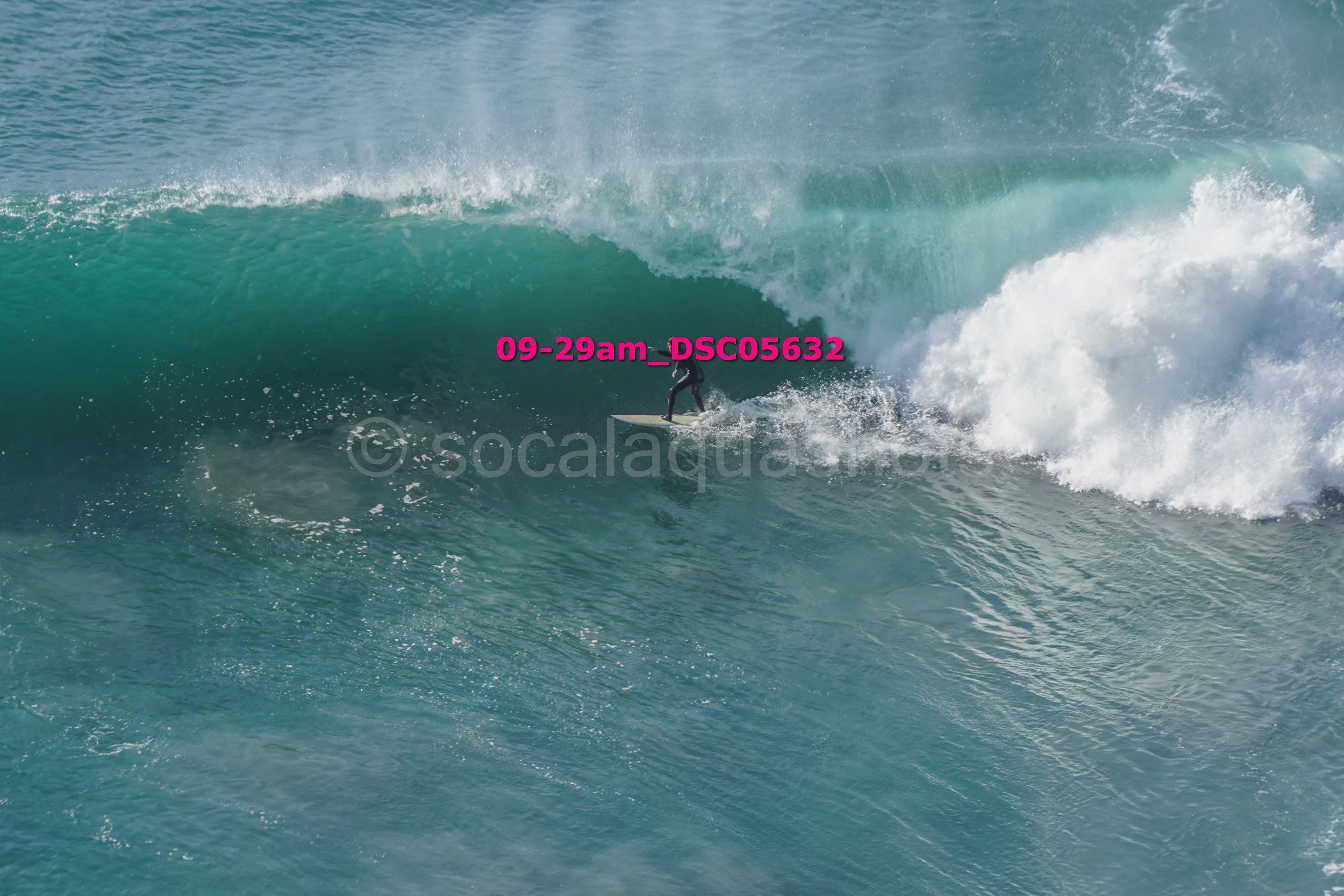 A person surfing on a large ocean wave with white foam, wearing a wetsuit, against a blue sky.