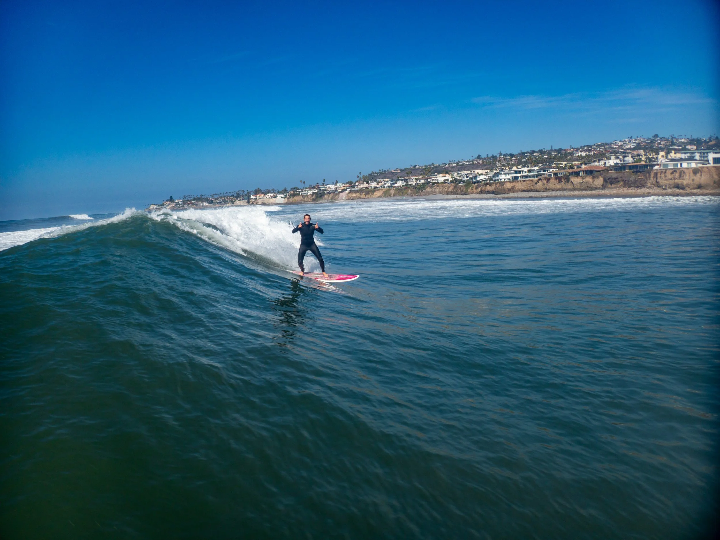 A person surfing on a wave with a coastal neighborhood in the background under a clear blue sky.