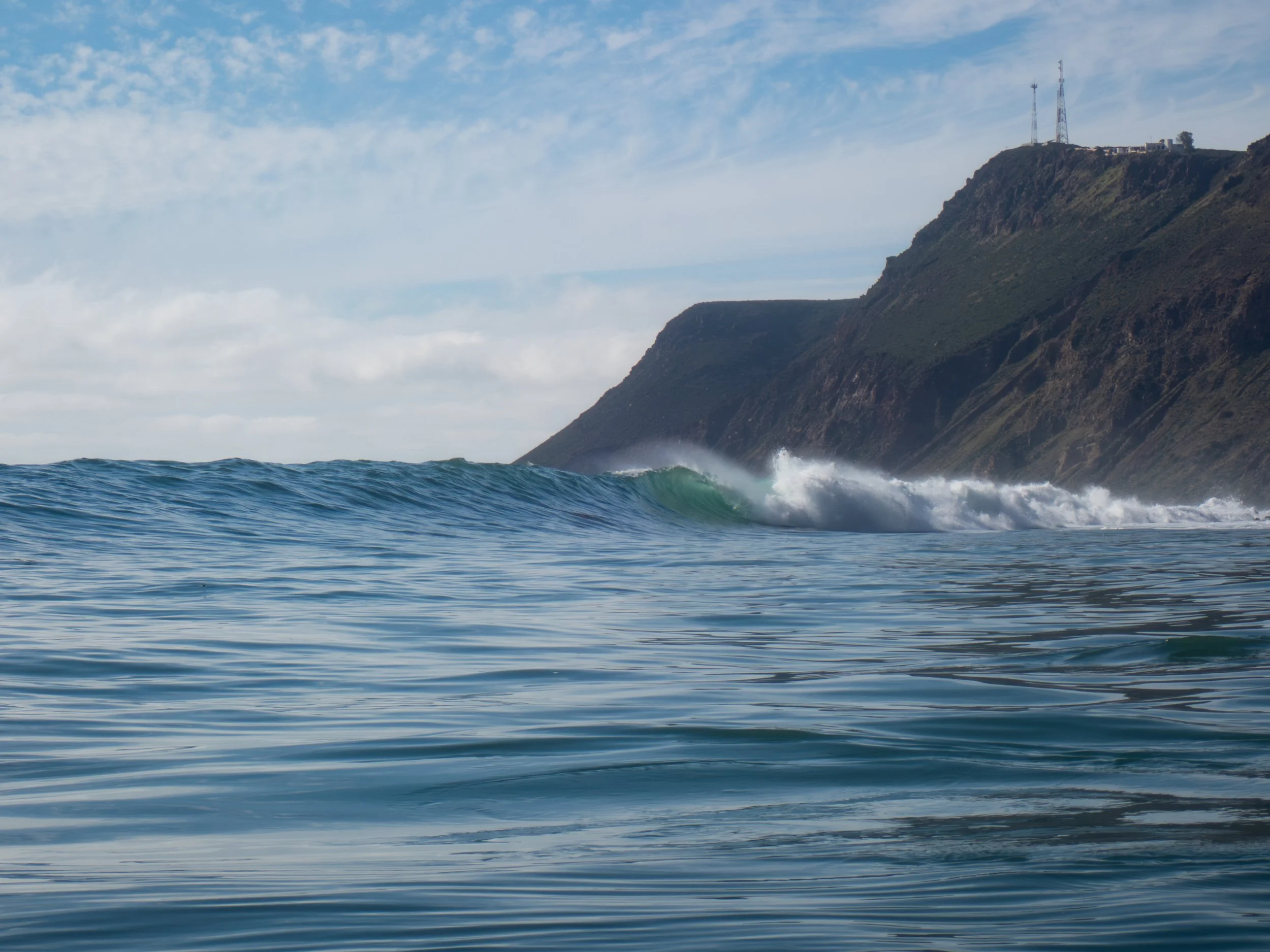 Ocean waves near a coastline with steep cliffs and communication towers on a cloudy day.