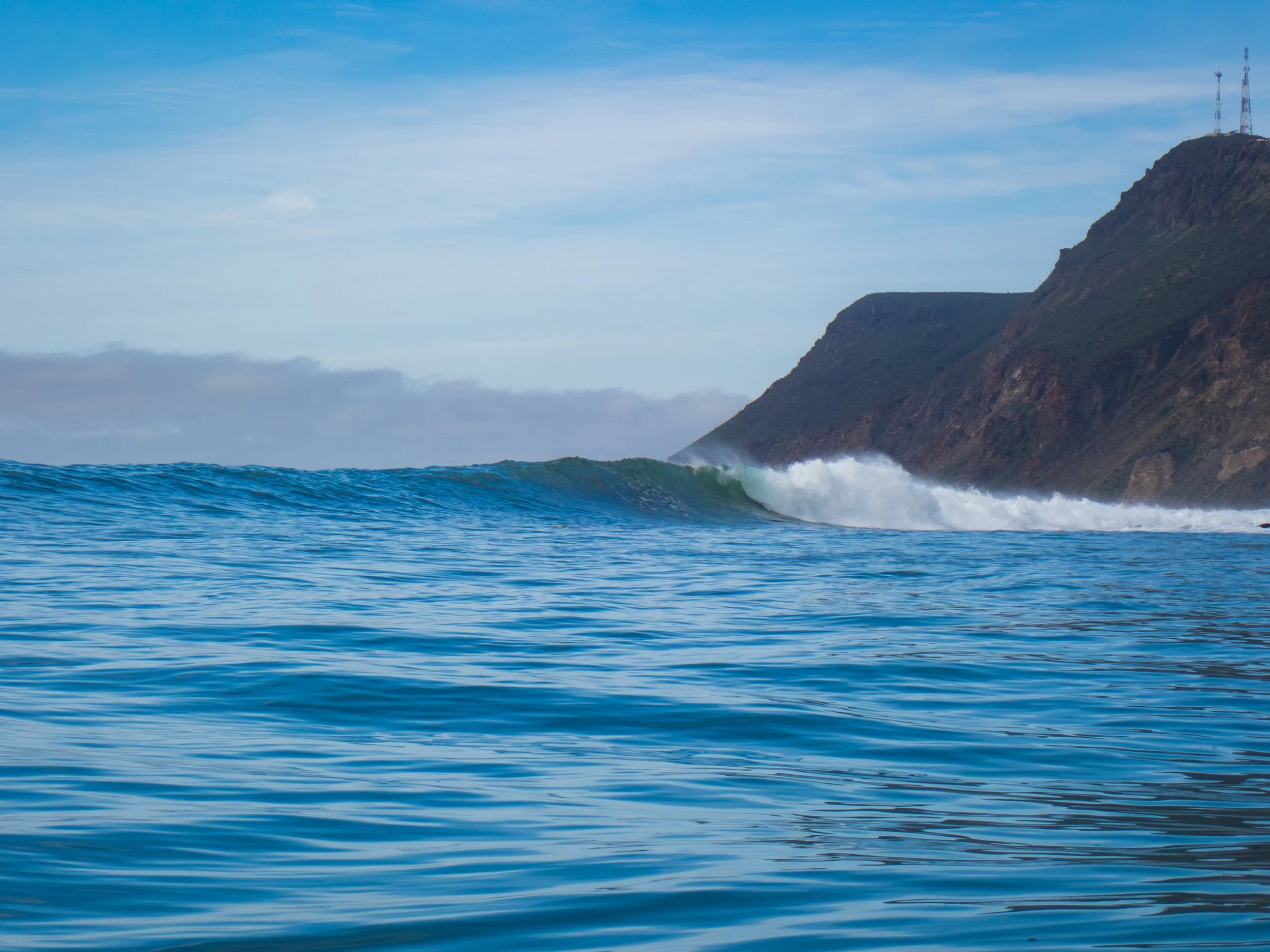 Vast ocean with a breaking wave near a mountainous coastline under partly cloudy skies.