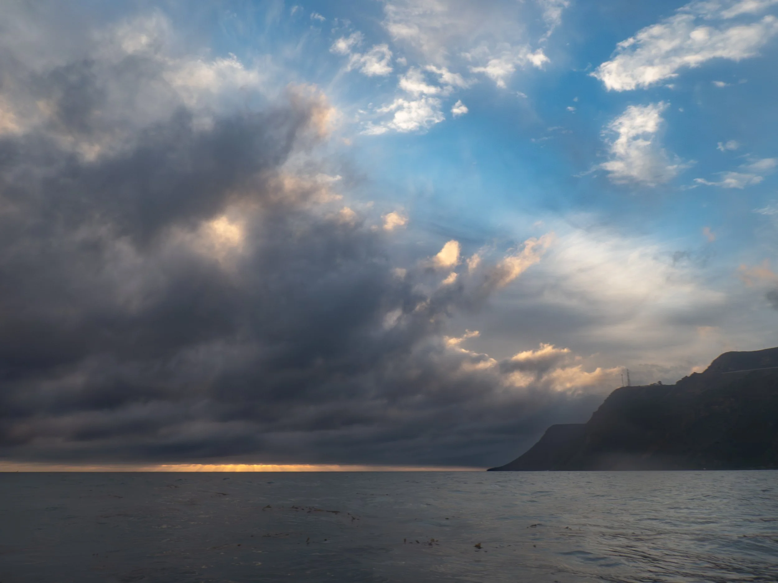 Overcast sky with dark and light clouds over a body of water, with a mountain or cliff on the right side of the horizon.