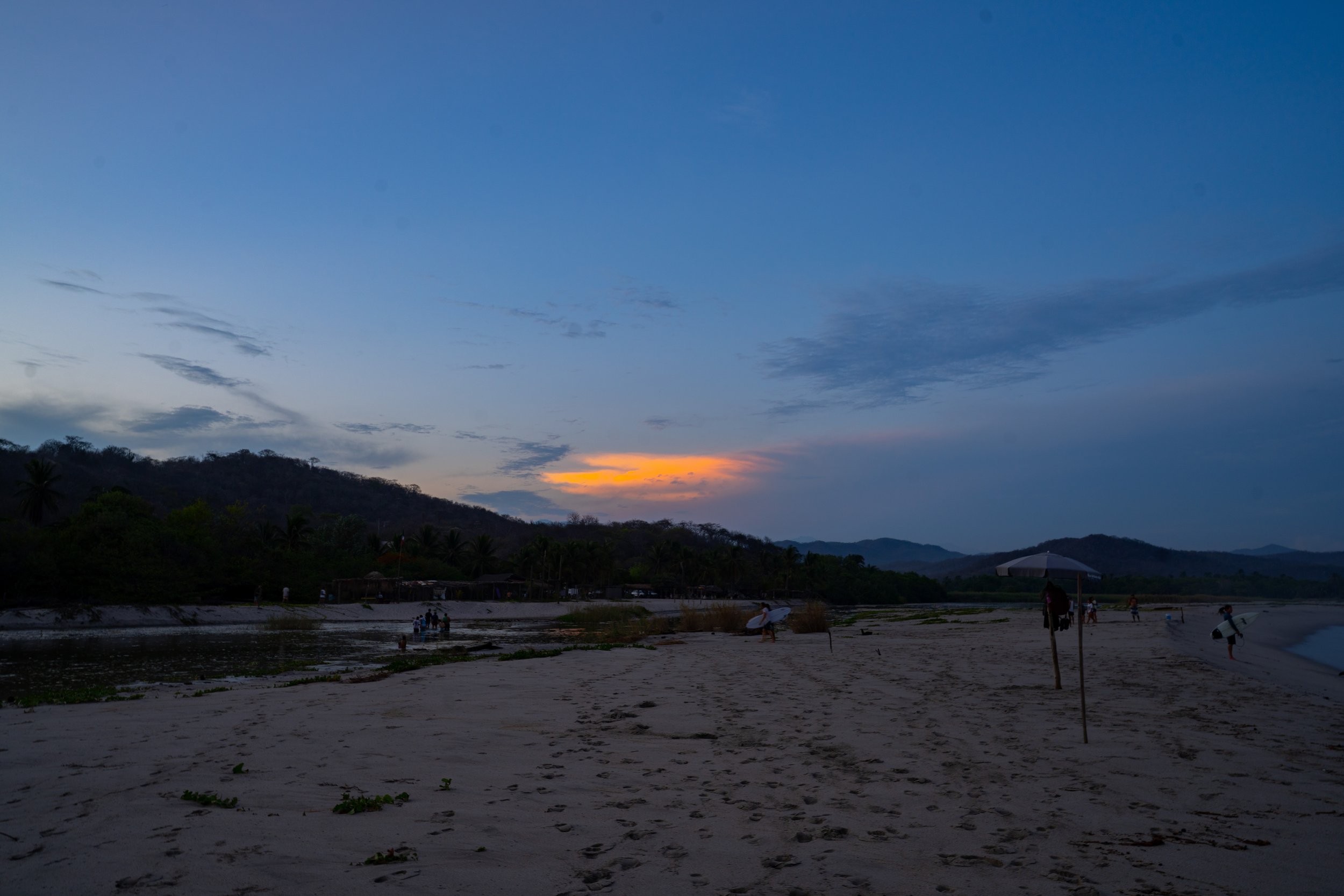 A beach scene at sunset with a partly cloudy sky, people walking and surfing, some hills in the background, and the horizon showing the sunset.