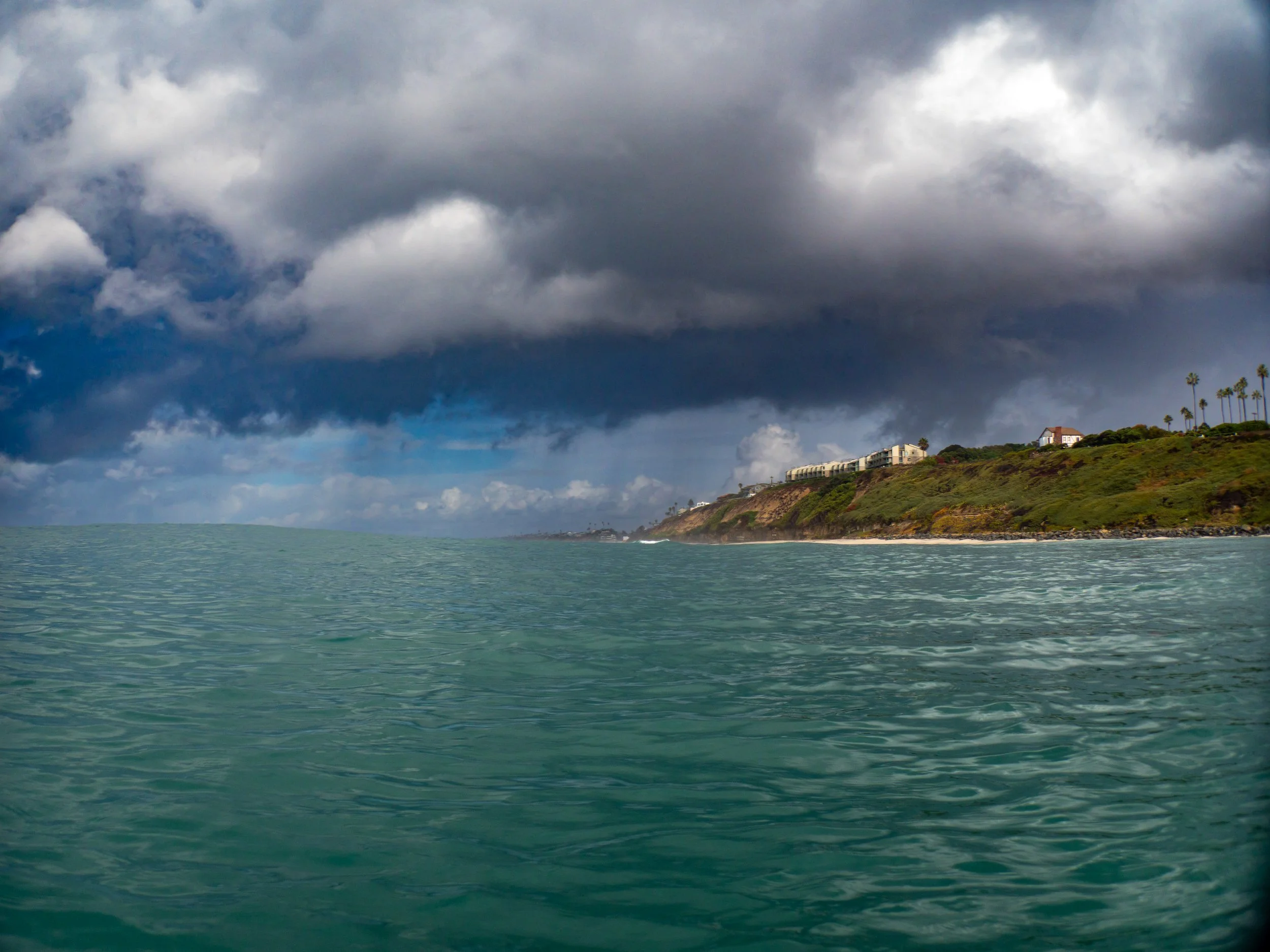 Ocean view with a cloudy sky and a hillside with houses and palm trees in the distance.