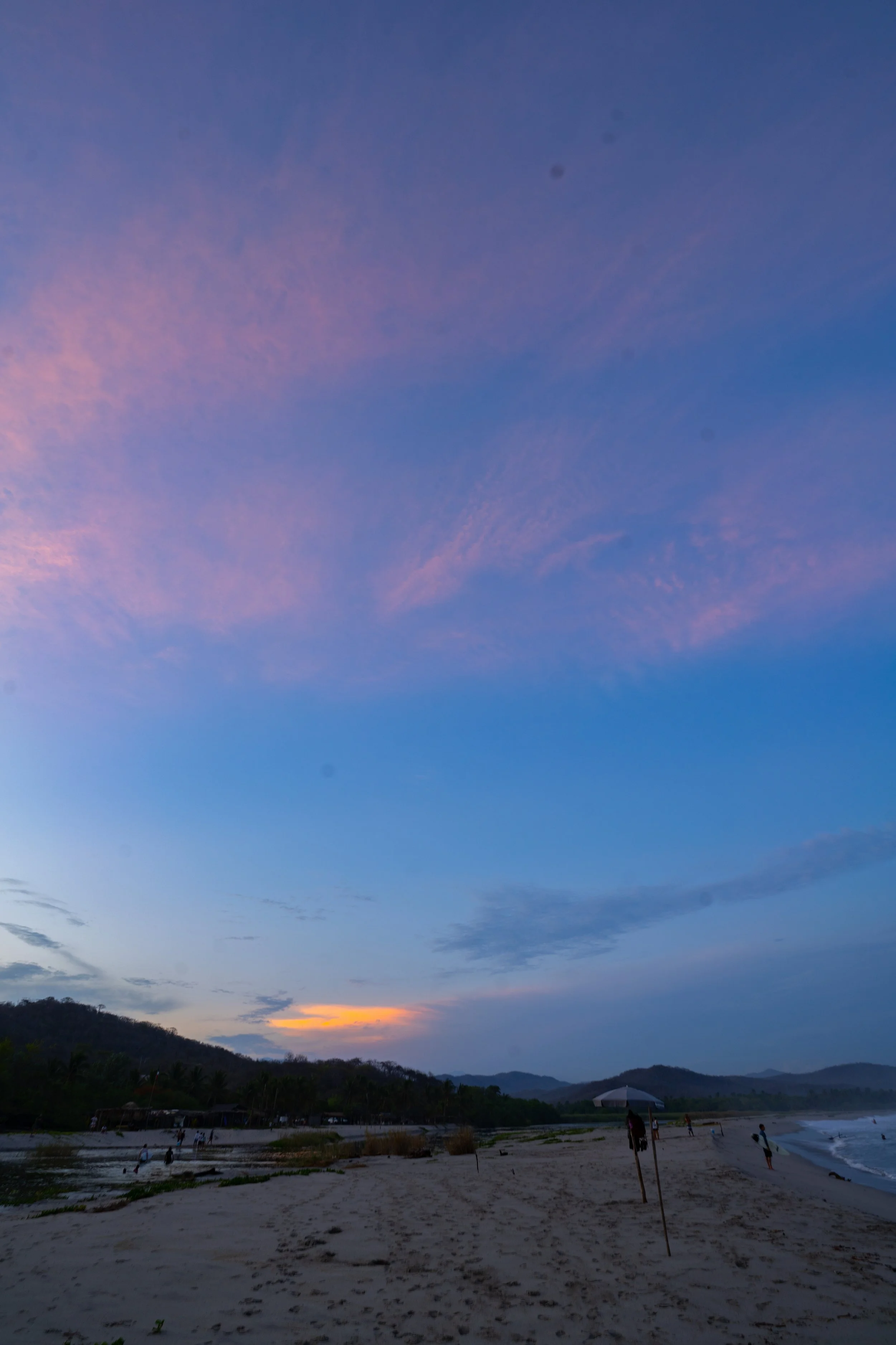 Beach at sunset with a cloudy sky and distant mountains, people walking along the shoreline.