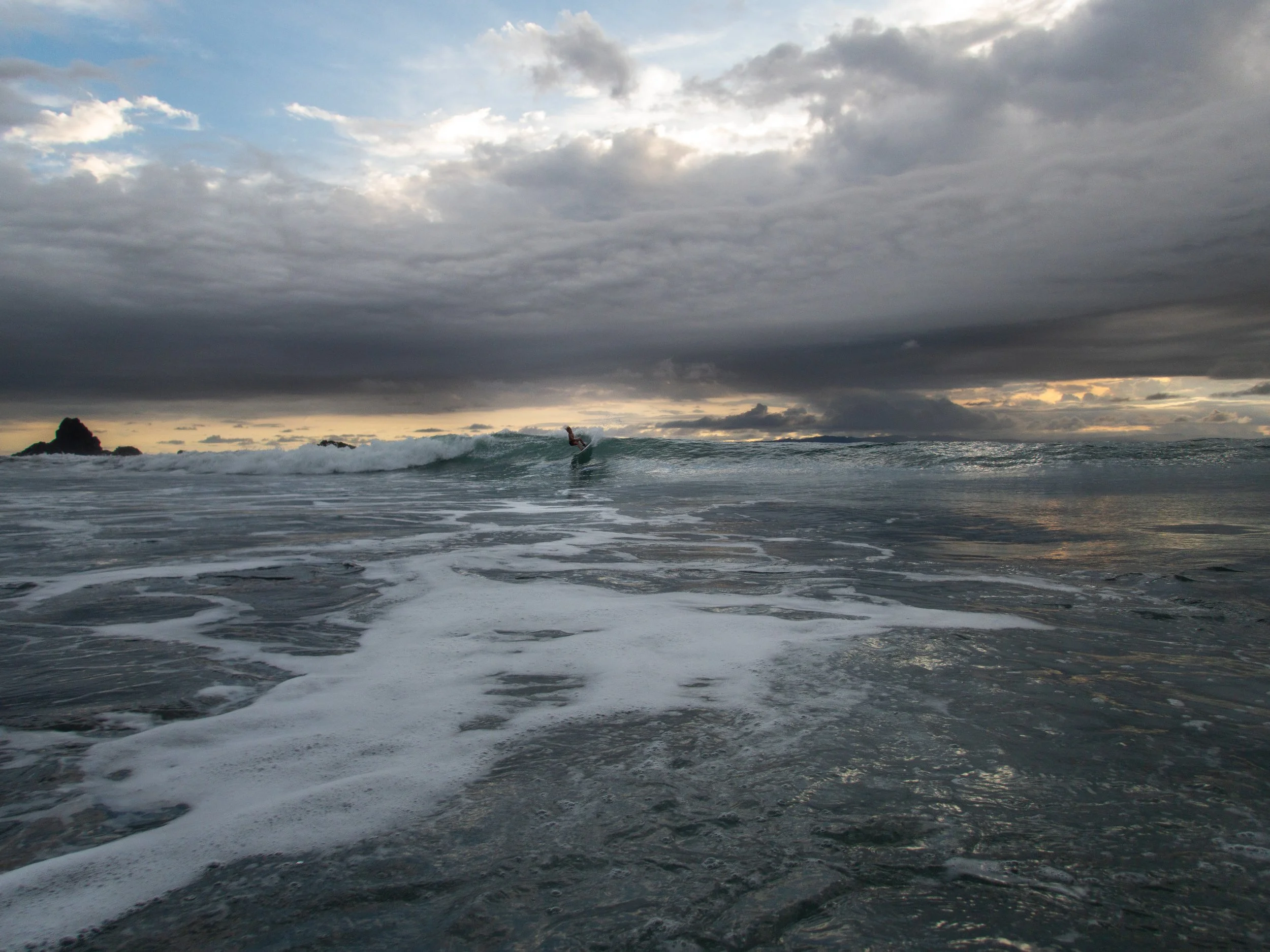 Surfer riding a wave under a cloudy sky at sunset, ocean scene with foam on the water.