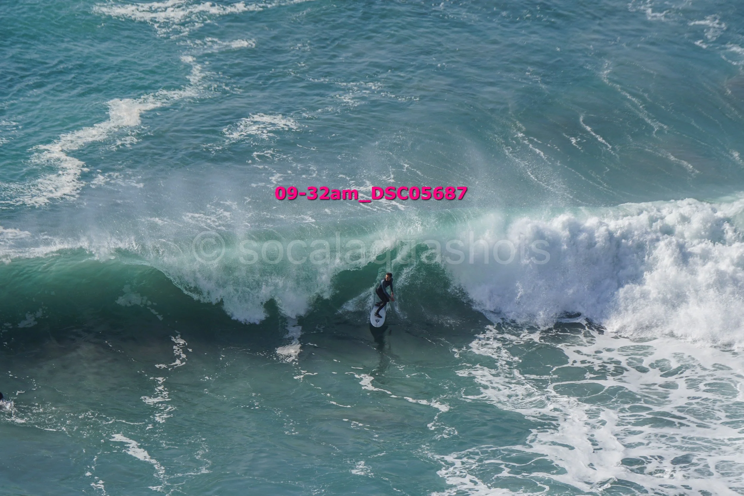 A person surfing on a large ocean wave with blue water and white foamy surf, wearing a wetsuit.