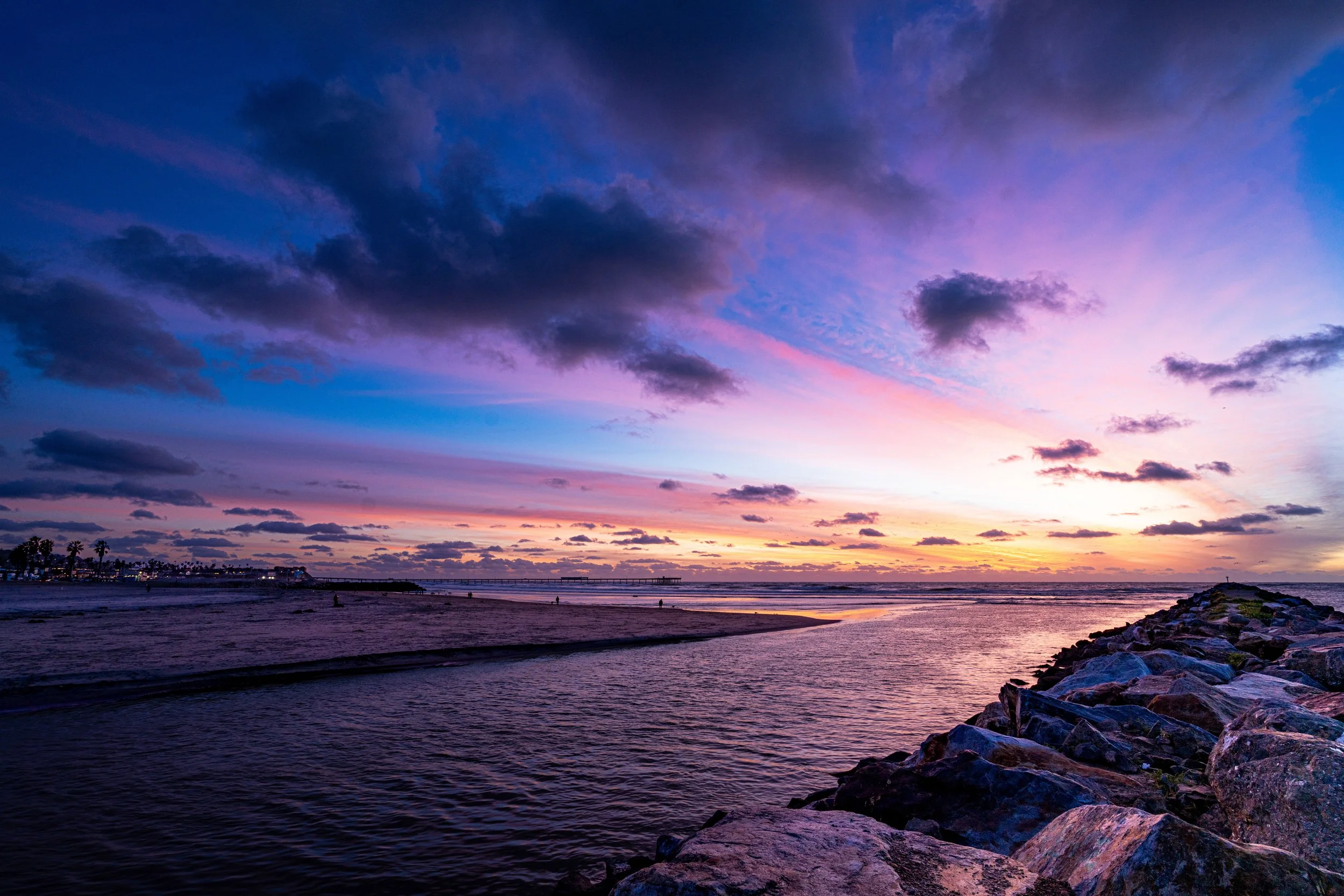 Colorful sunset over a beach with rocks and a pier in the distance.