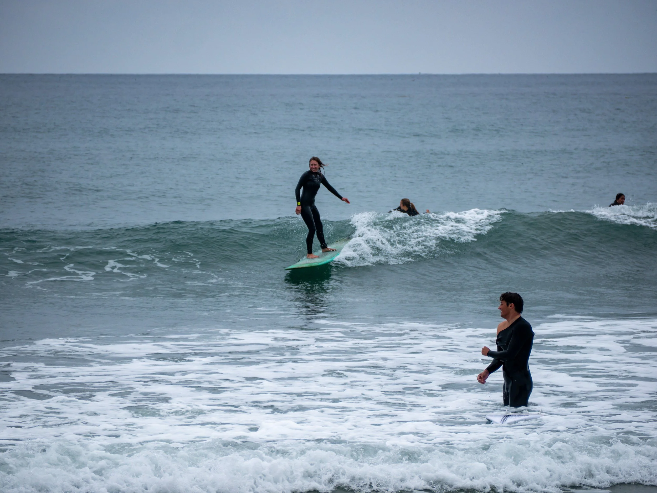 Person surfing on a wave with other surfers and a person standing in the water nearby.