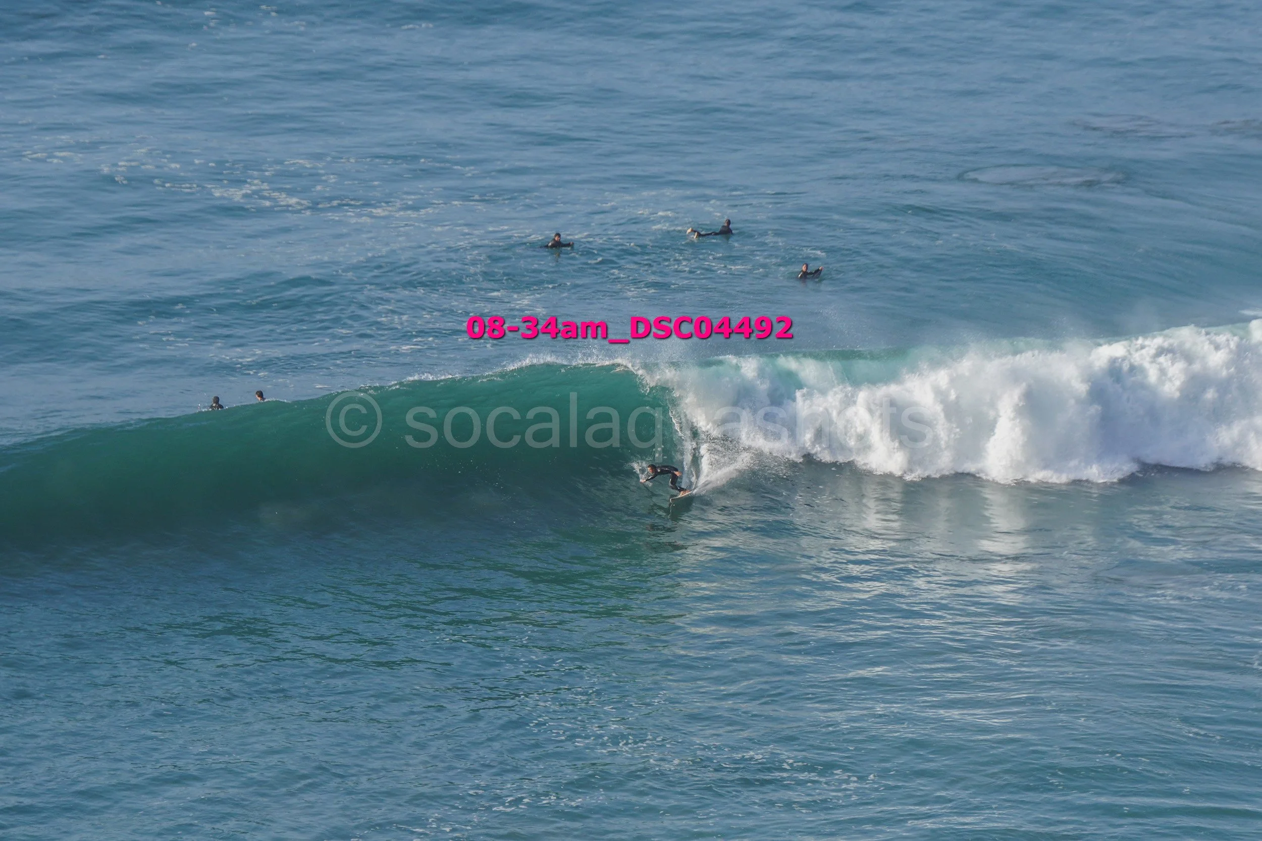 Surfer riding a wave in the ocean with several people floating on surfboards in the background.