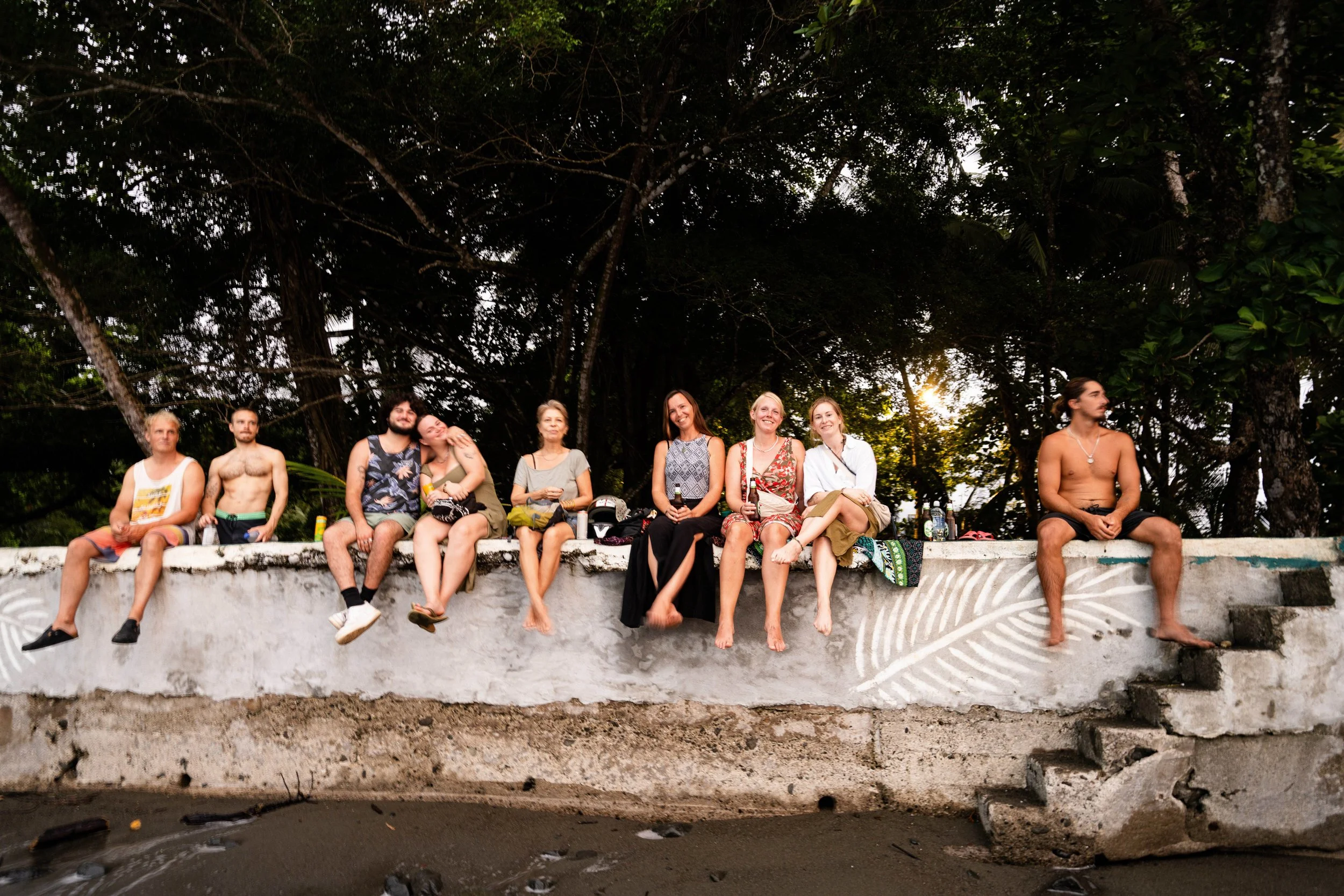Group of nine people sitting on a concrete ledge by a body of water, with dense trees in the background during sunset. The group includes men and women dressed in casual and summer clothes, some shirtless, holding drinks, and engaging in conversation