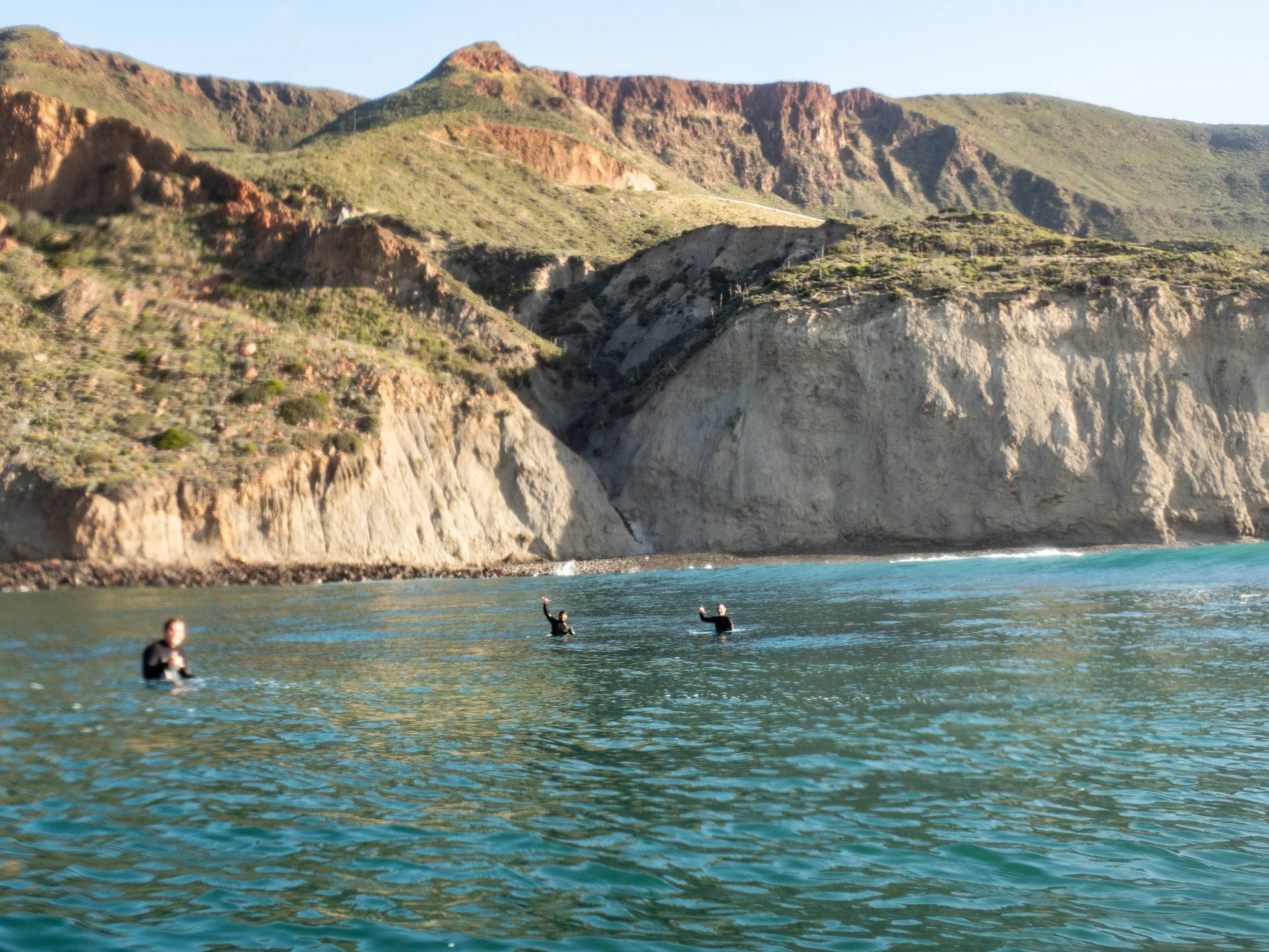 Three people swimming in ocean waters near rocky cliffs and mountainous terrain in the background.