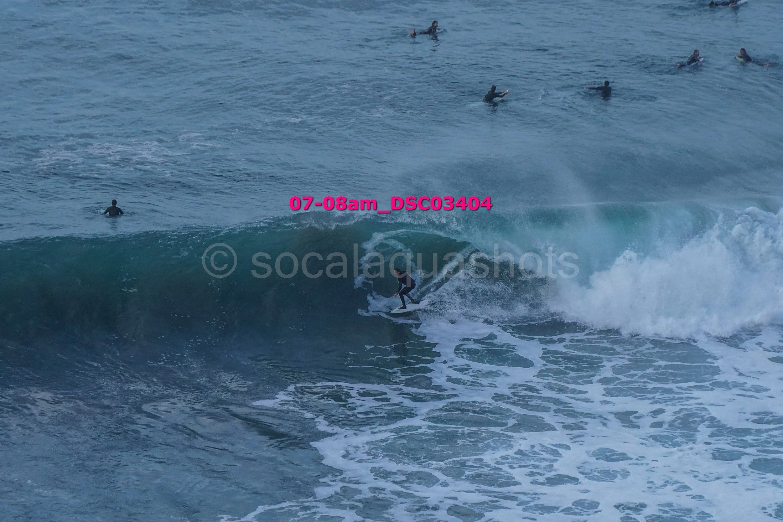 A surfer riding a wave with multiple people swimming and surfing in the water in the background.