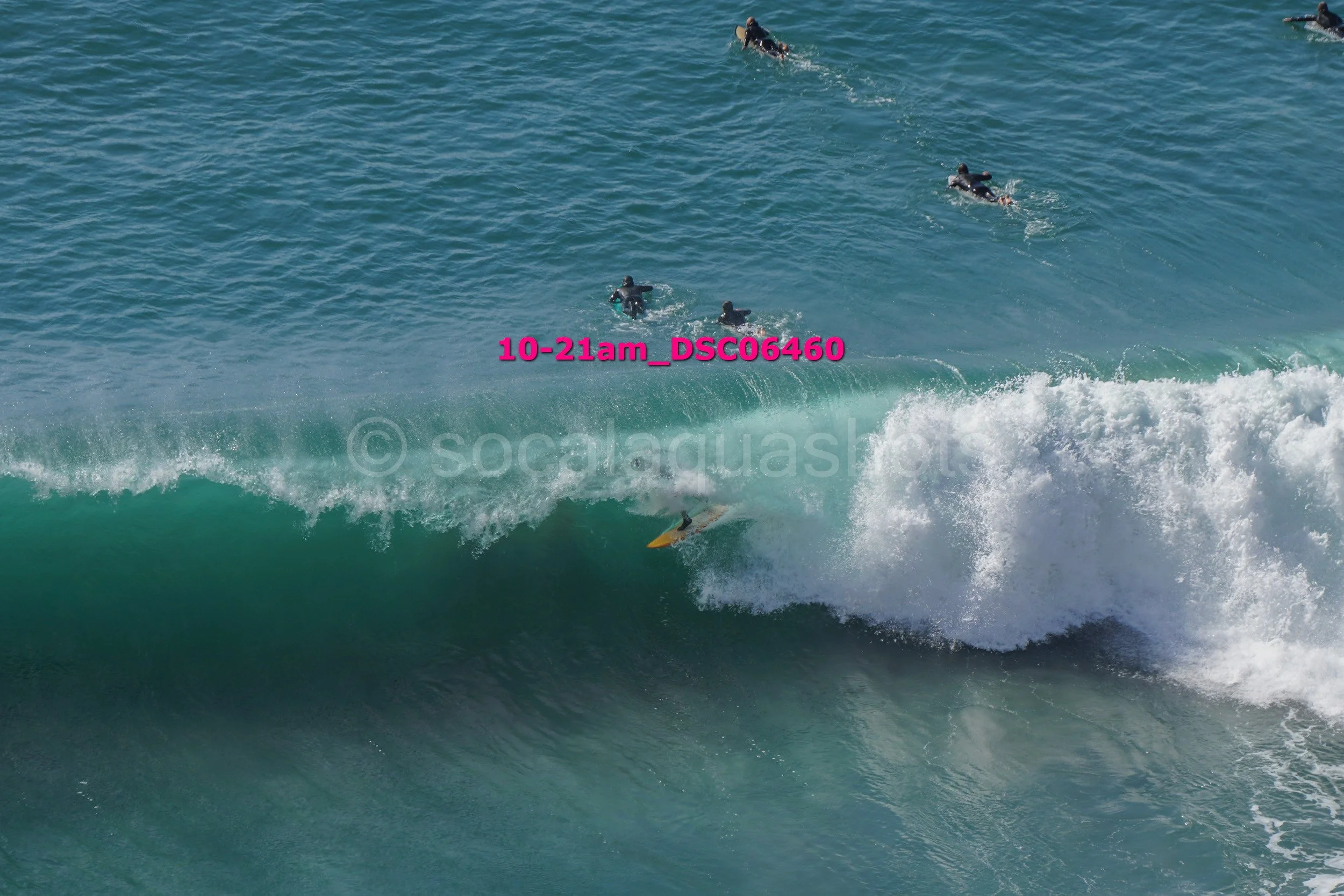 Surfer falling off a surfboard in a large breaking wave as surfers float in the water nearby.