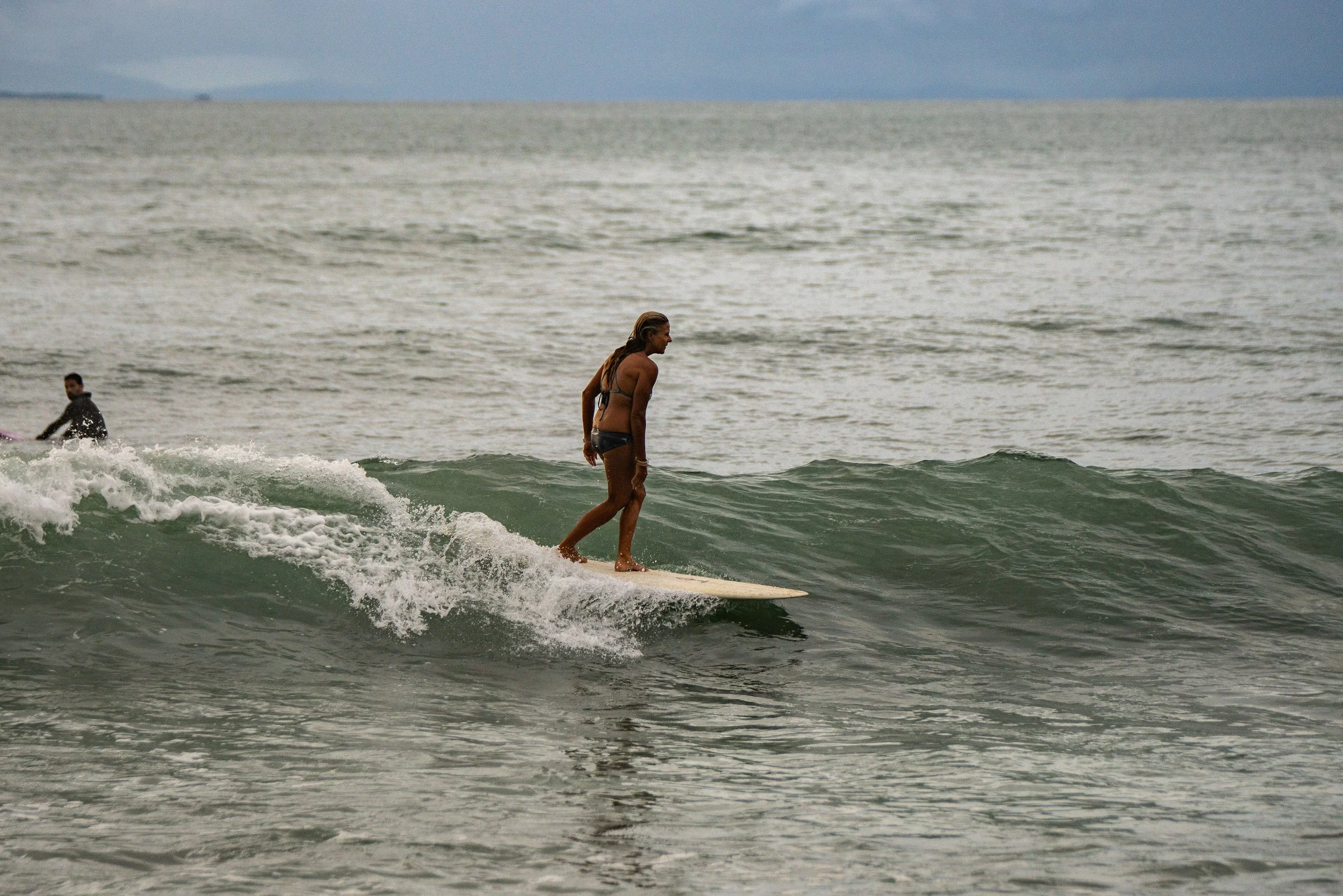 A woman surfing on a surfboard in the ocean, with a person in the background, under cloudy skies.