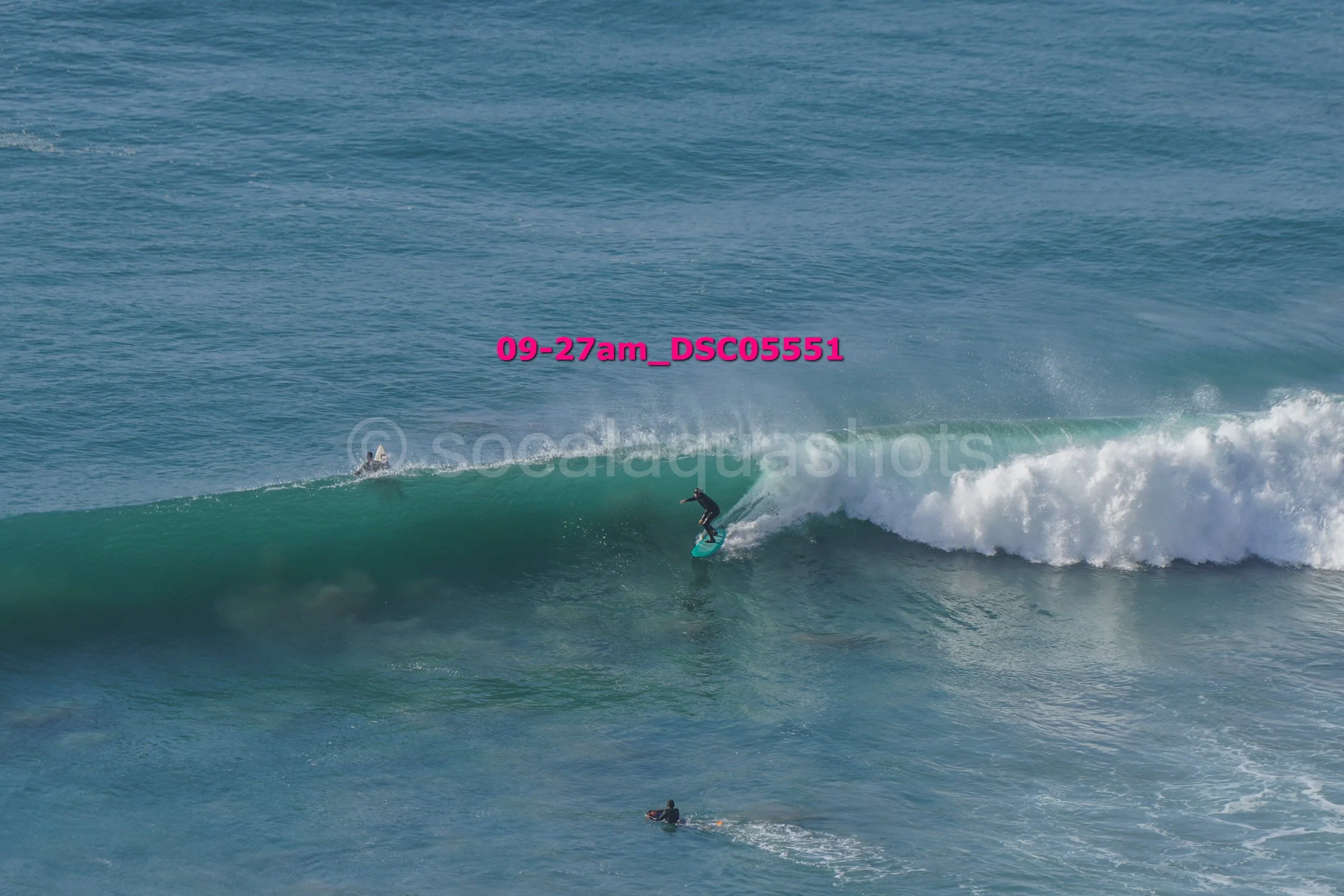 A person surfing a large wave in the ocean, with other surfers visible in the water.