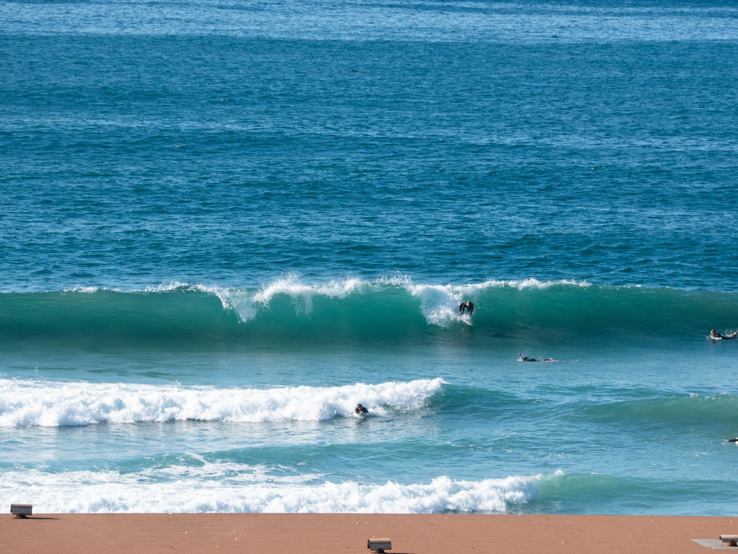 Surfers riding waves at the beach with clear blue water and sandy shore.