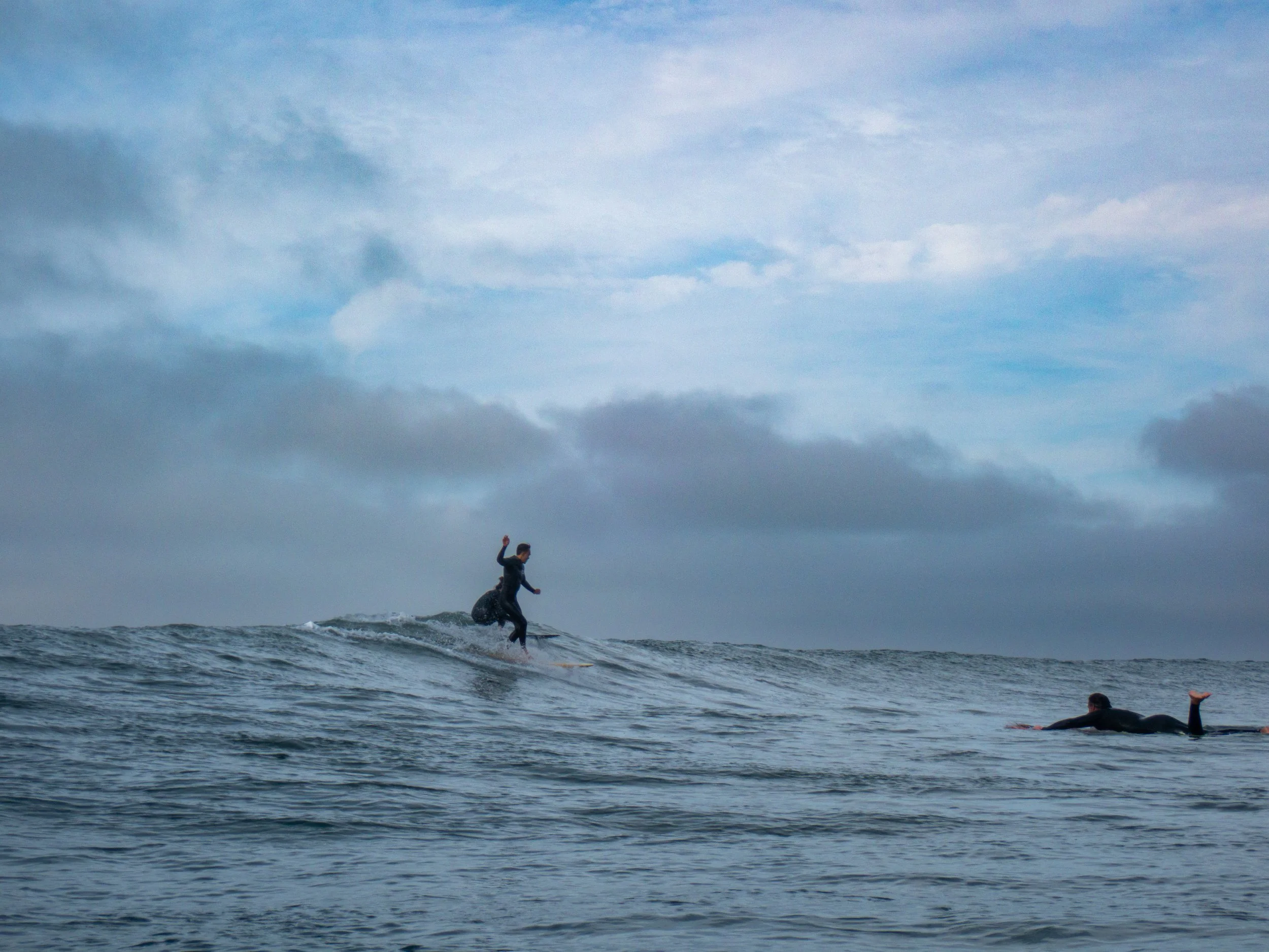 One person surfing a wave and another swimming in the water under a cloudy sky.