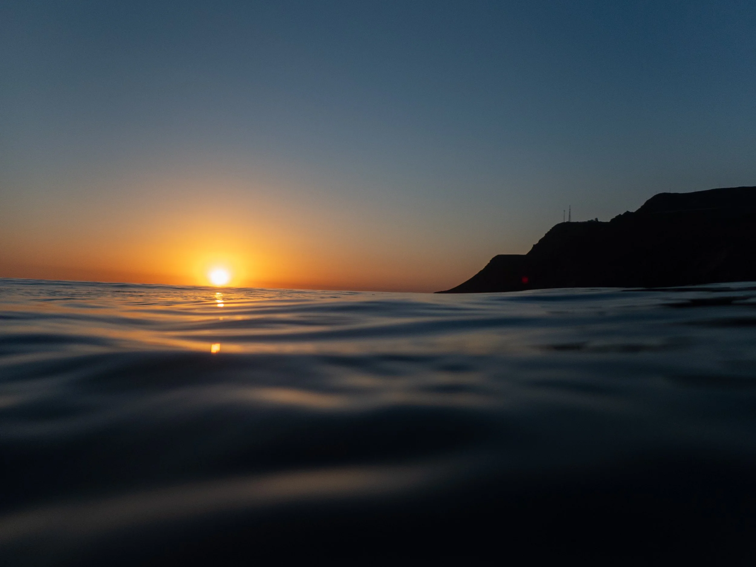 Sunset over the ocean with a dark hillside on the right and calm water in the foreground