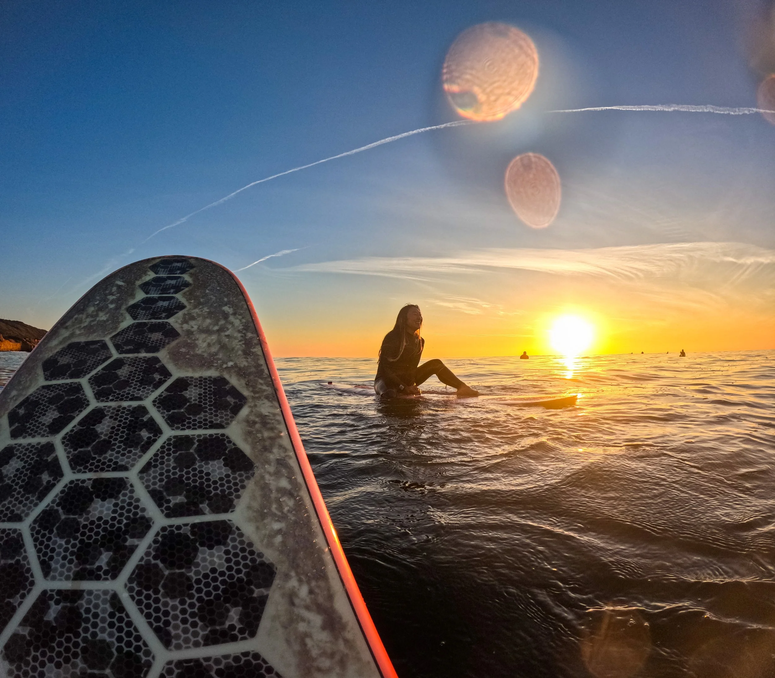 A person sitting on a surfboard in the water at sunset, with the sunset in the background and a surfboard in the foreground.