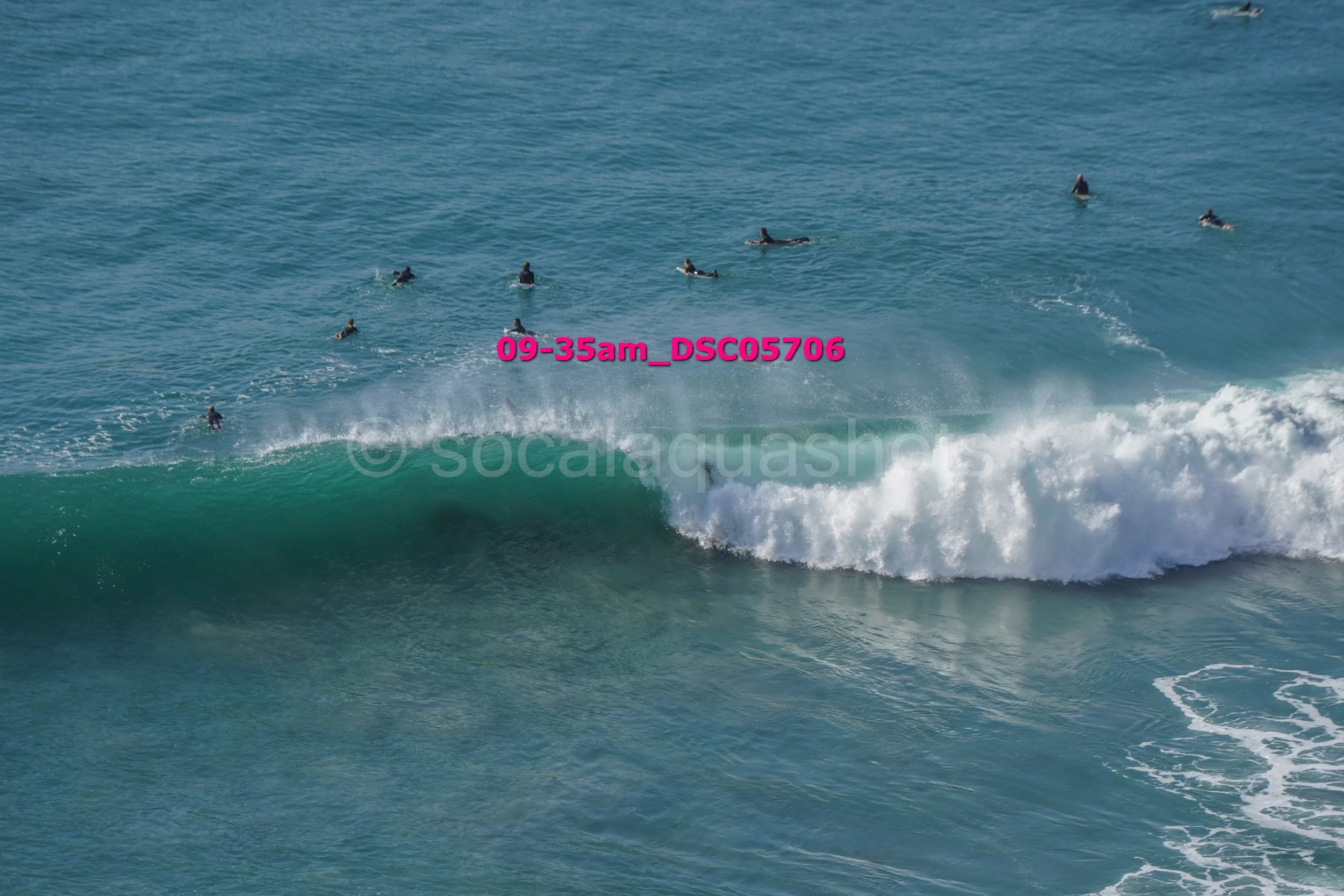 Multiple surfers in the ocean waiting for waves as one surfer catches a wave, with water spray and white foam, under a clear sky.