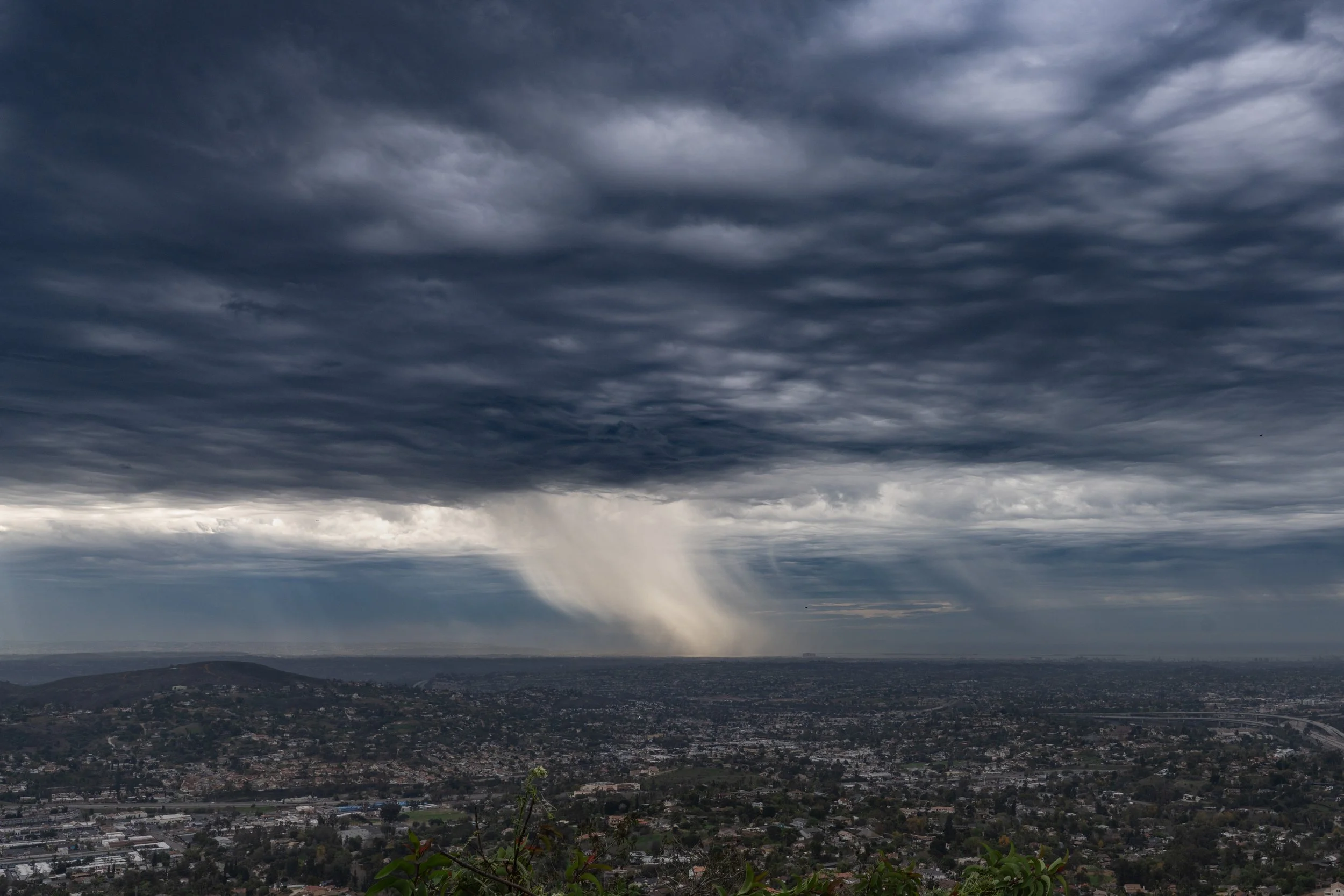 Dark, stormy clouds with rain falling over a cityscape and hills in the background.